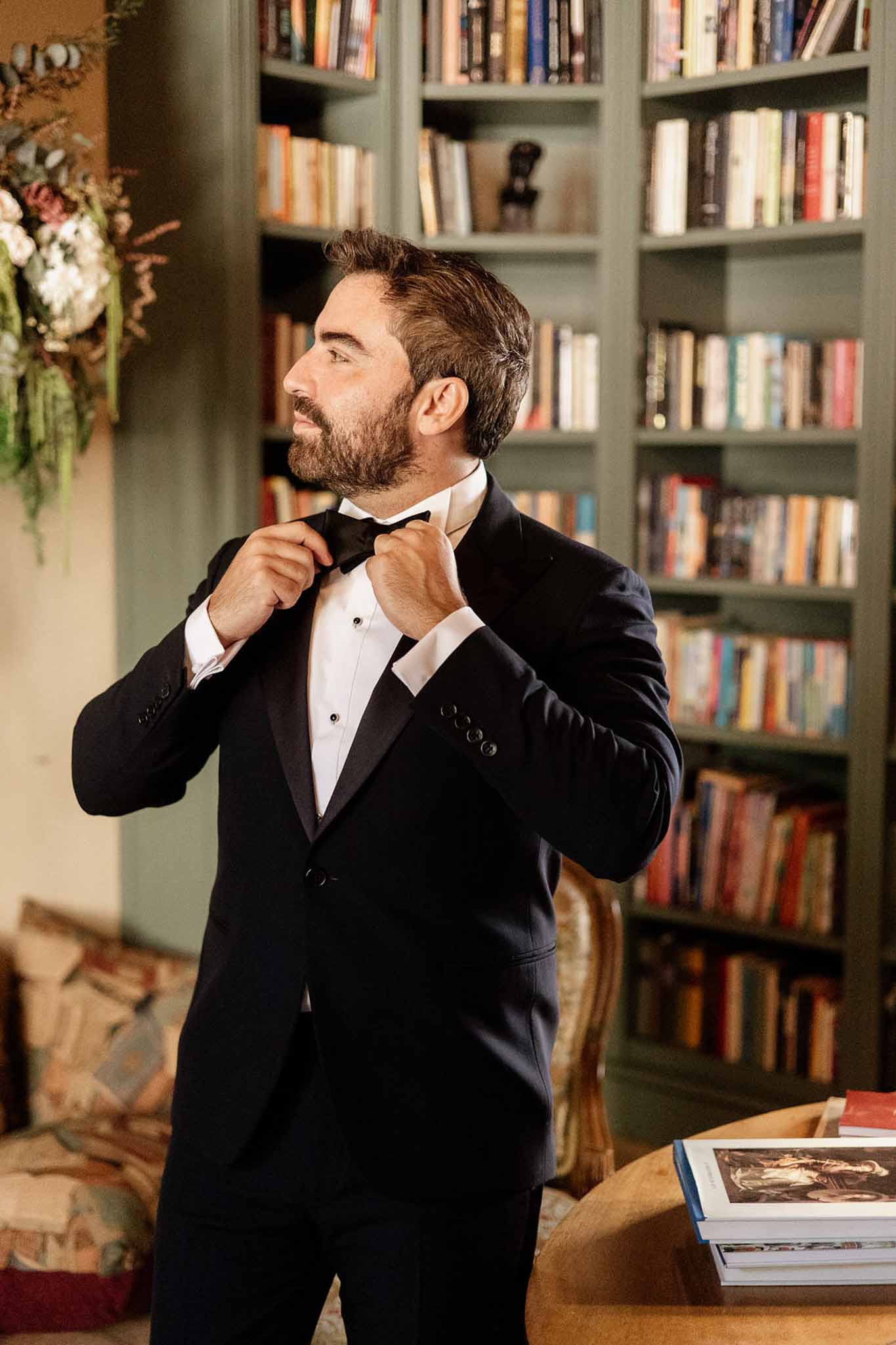 Groom adjusting black bow tie in sage green library with floor-to-ceiling bookshelves and patterned armchair