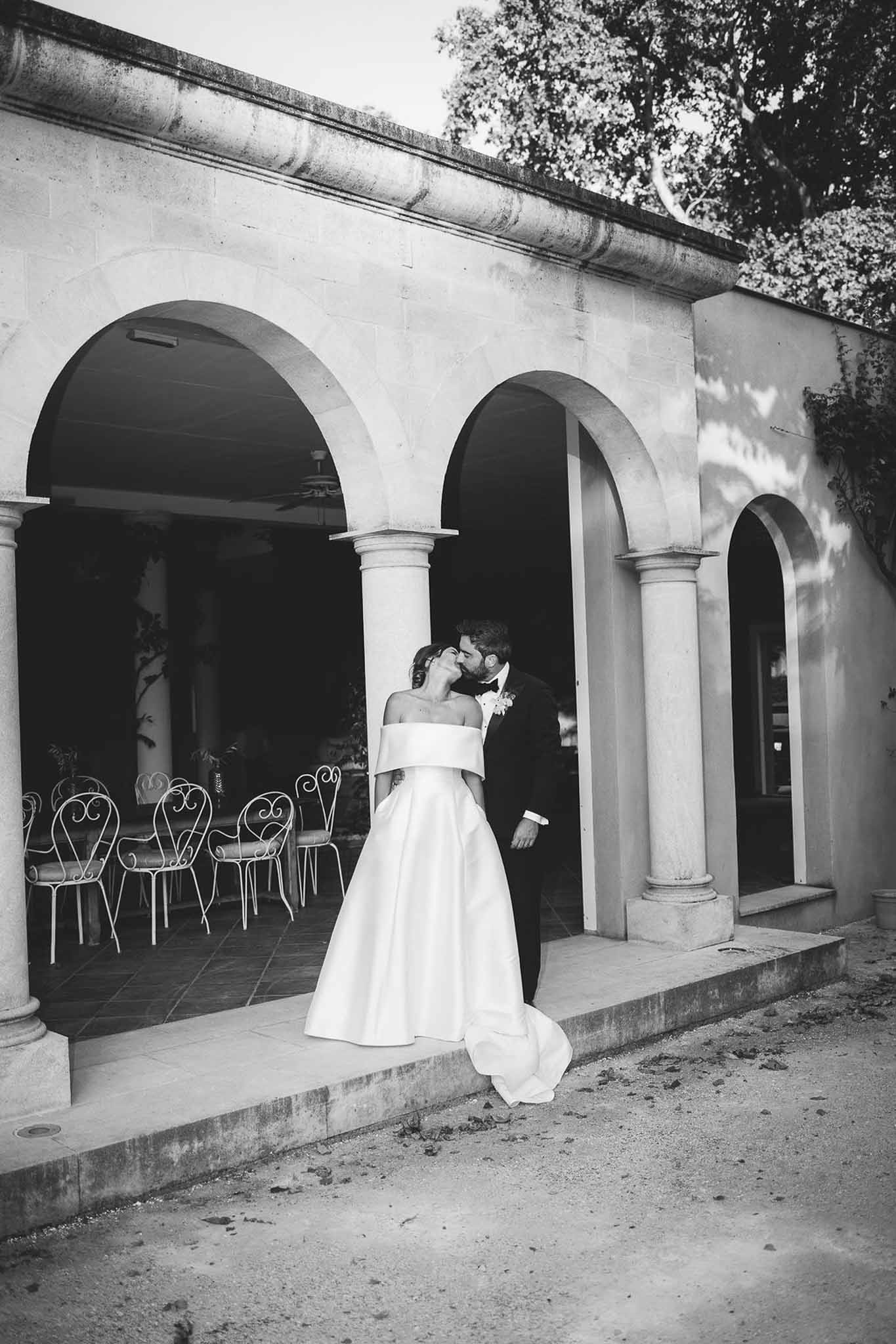 Black-and-white portrait of bride and groom kissing beneath stone archway colonnade at a French chateau