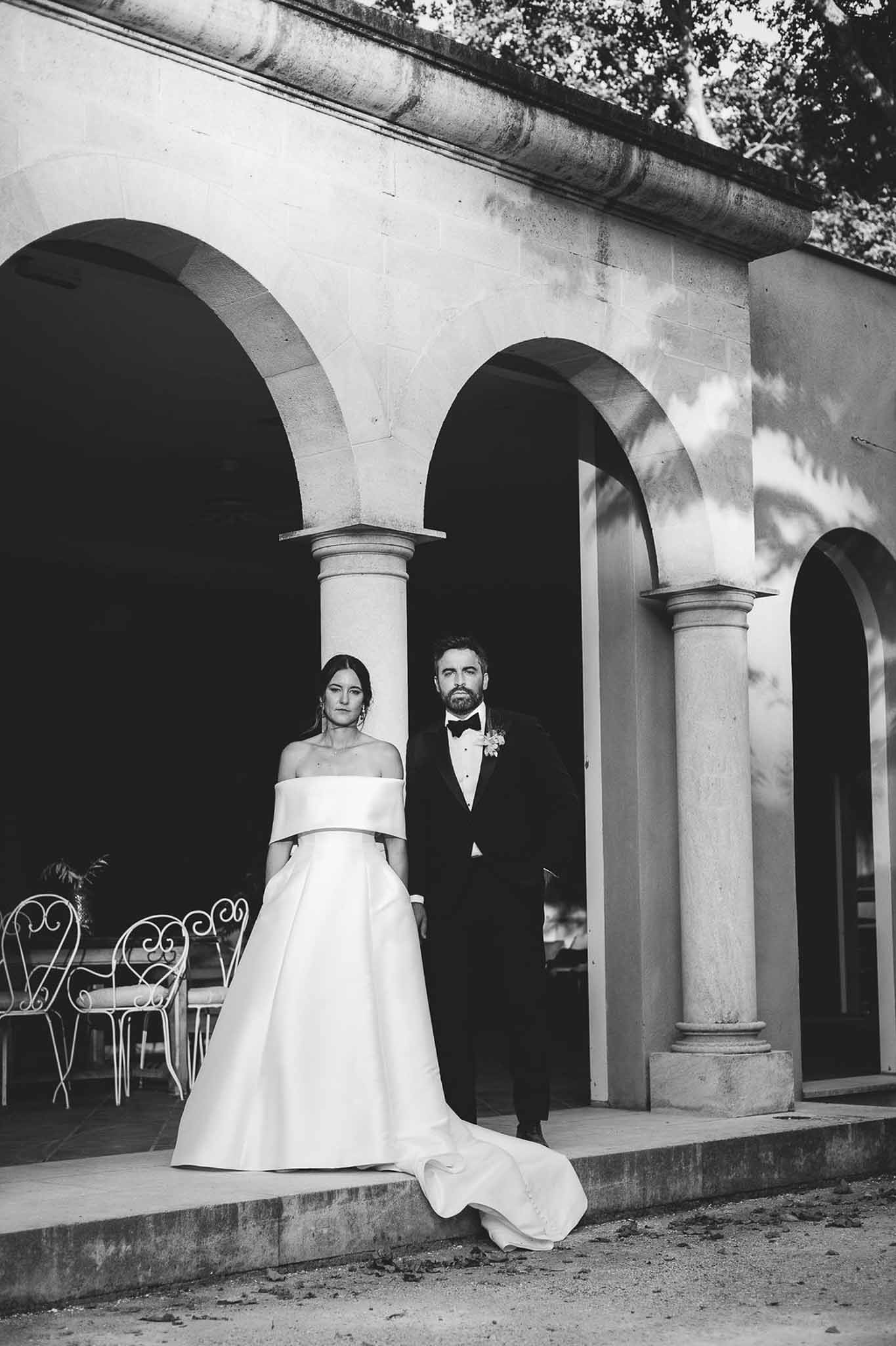 Black and white couple on stone steps beneath arched colonnade with wrought-iron garden furniture beyond