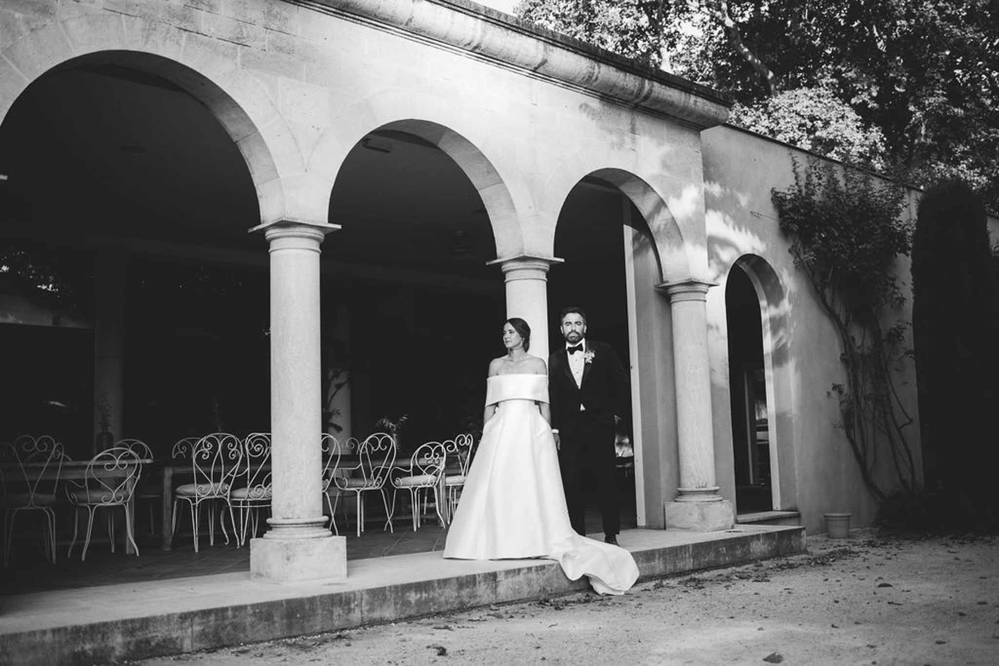 Black and white couple between stone columns with arched arcade and reception tables behind