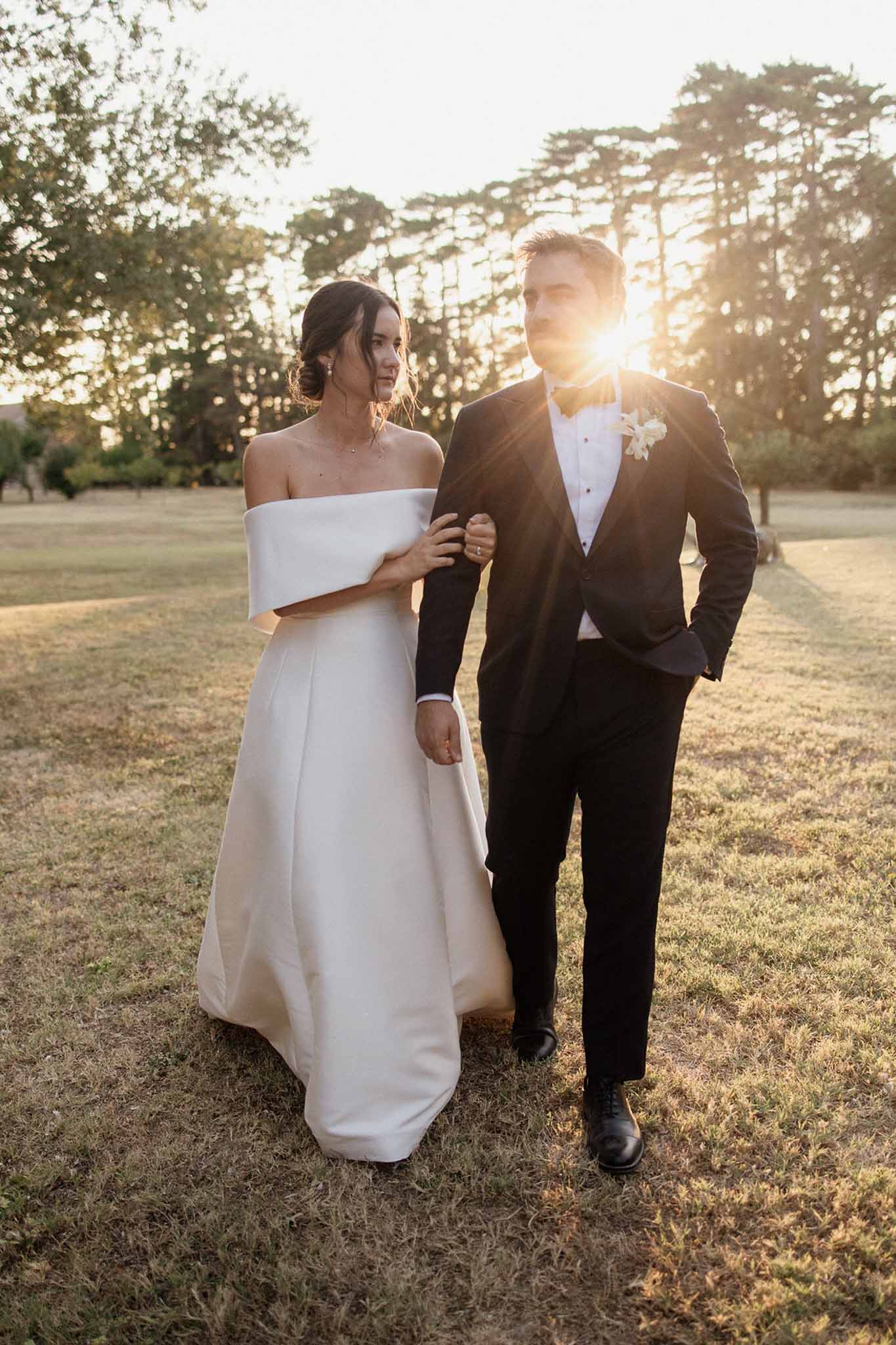 Bride in off-shoulder ivory ballgown and groom in black tuxedo walking arm in arm at golden hour