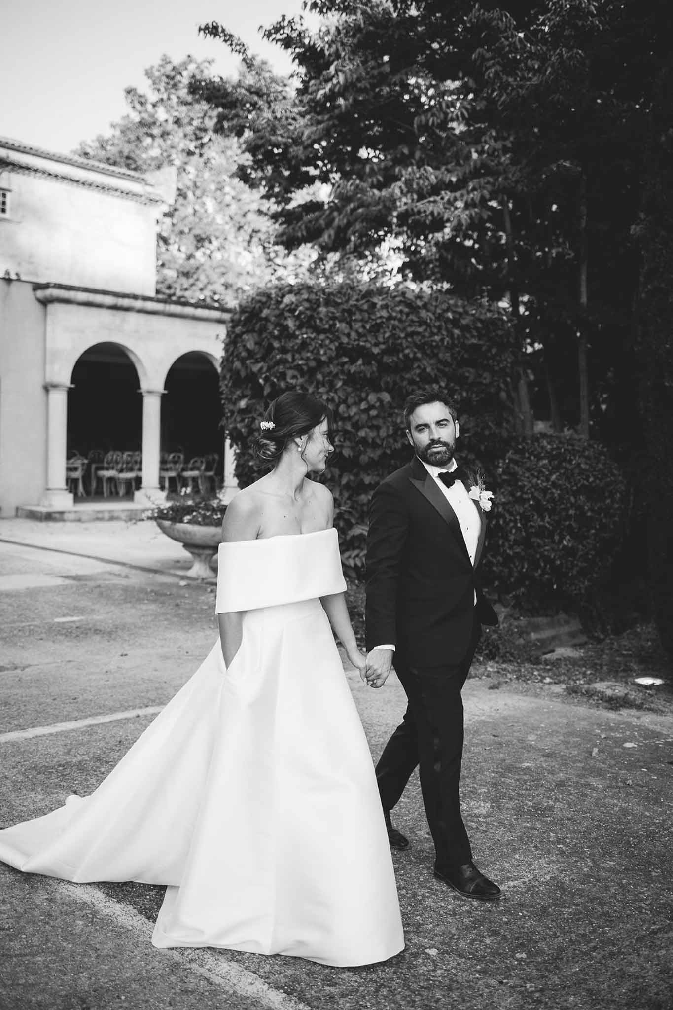 Black-and-white portrait of bride and groom walking hand in hand on chateau grounds with stone loggia behind