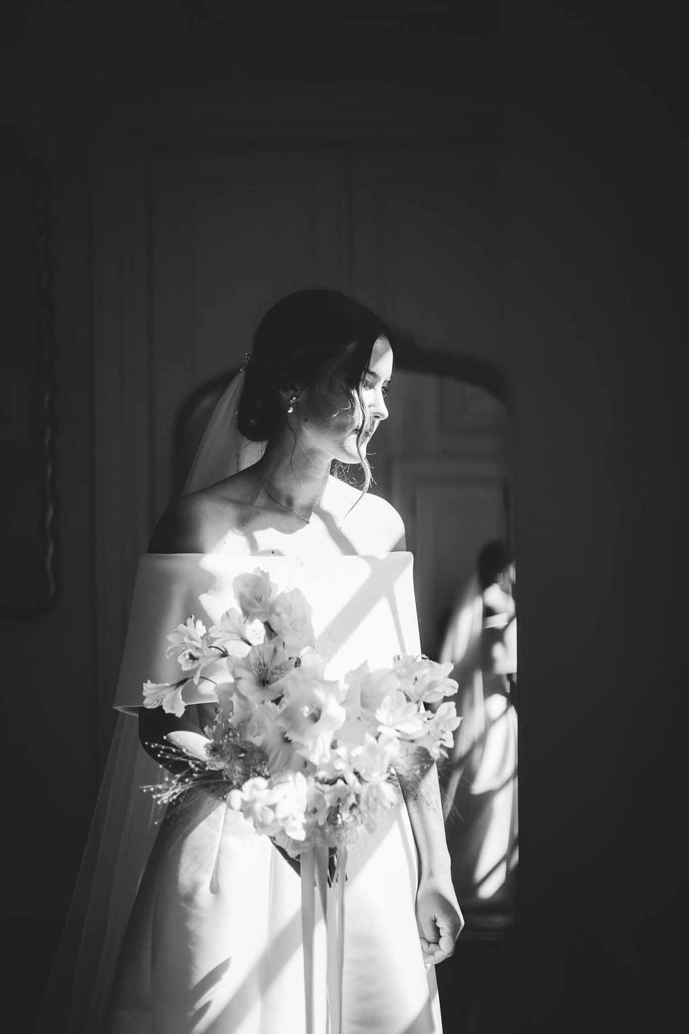 Bride laughing in shaft of window light holding loose bouquet with dramatic shadow patterns in B&W