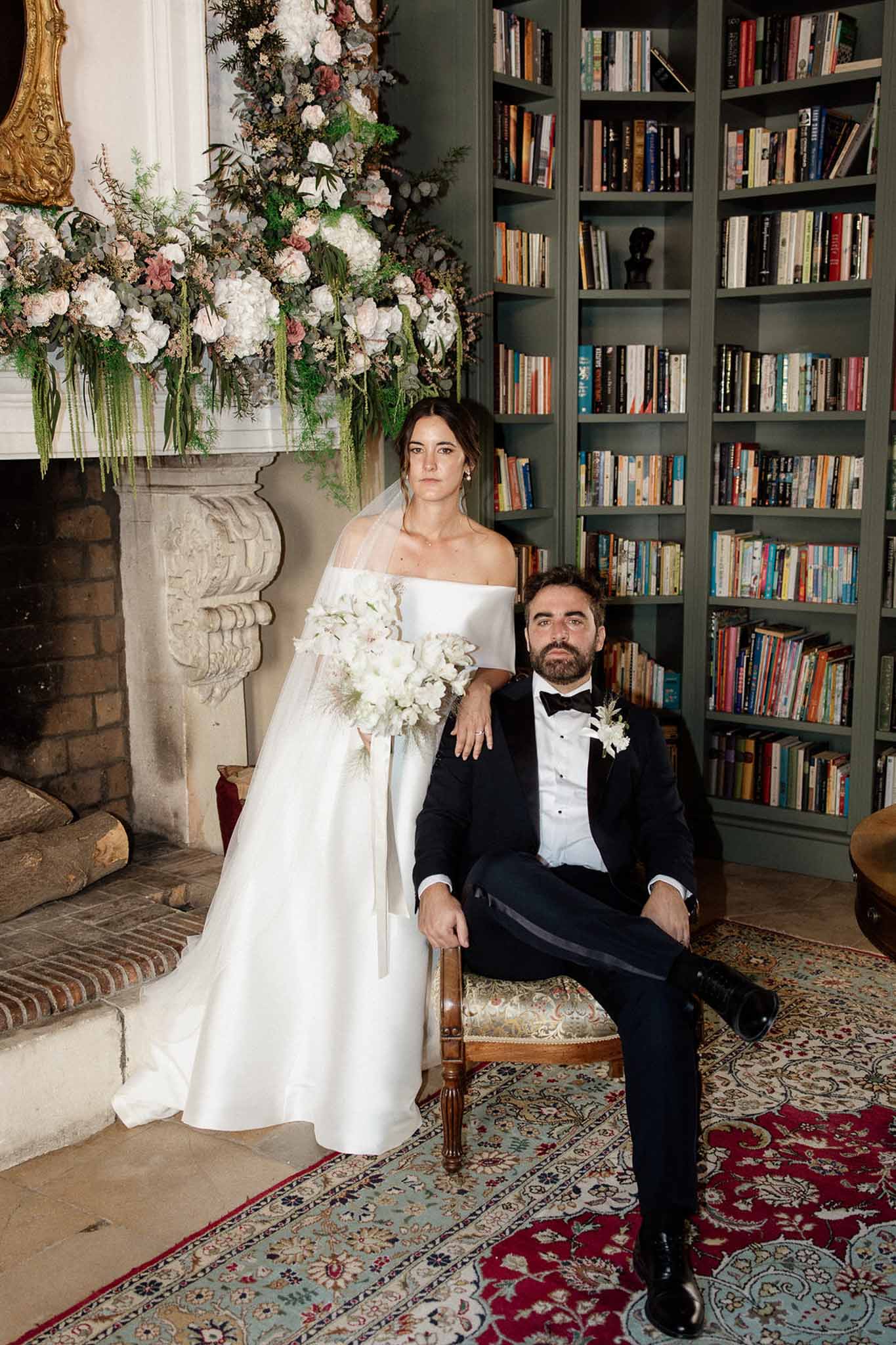 Bride in ivory ball gown and groom in black tuxedo posed in chateau library with floral-decorated fireplace mantel