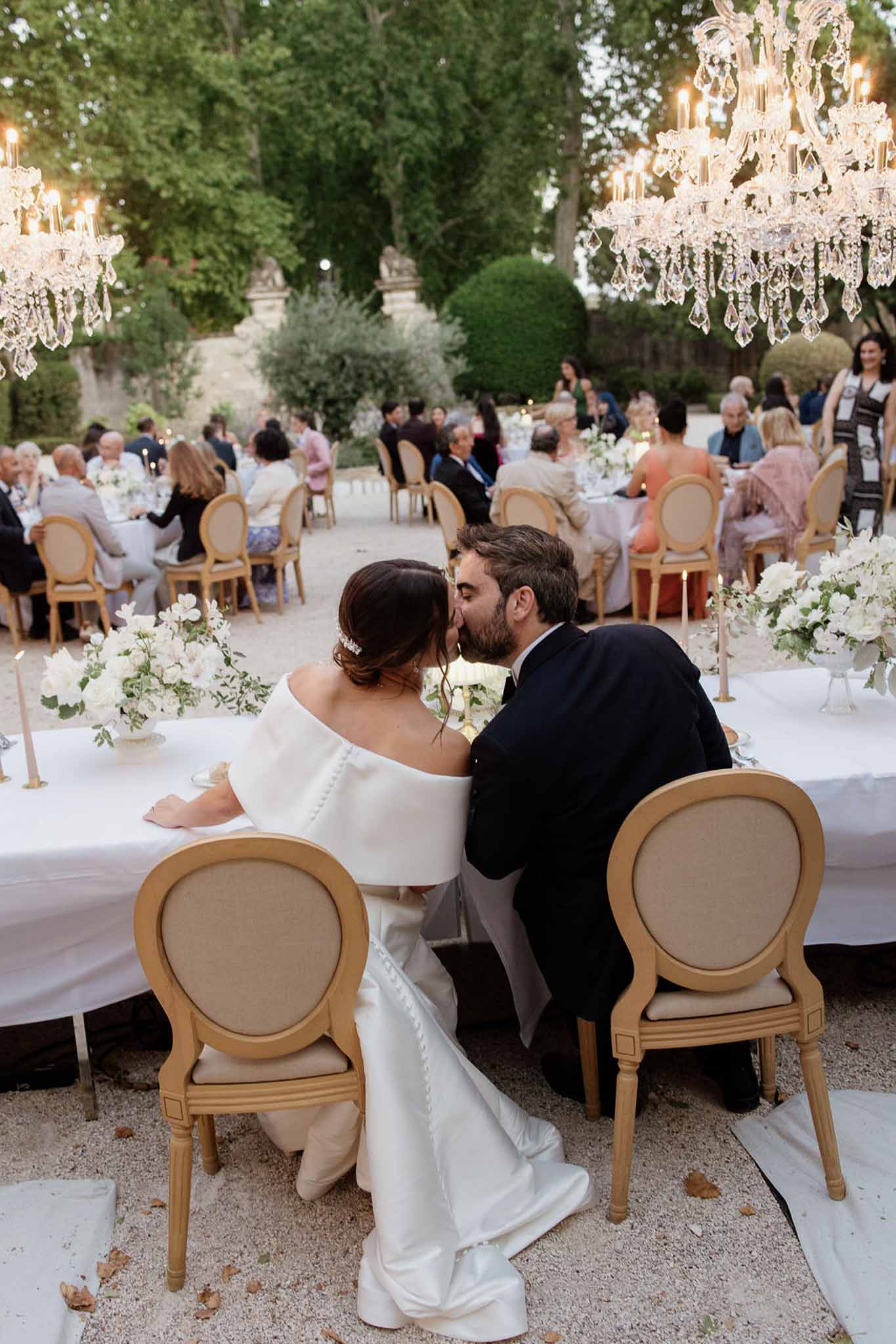 Couple kissing at sweetheart table with crystal chandeliers, white rose centrepieces, and 50 guests behind