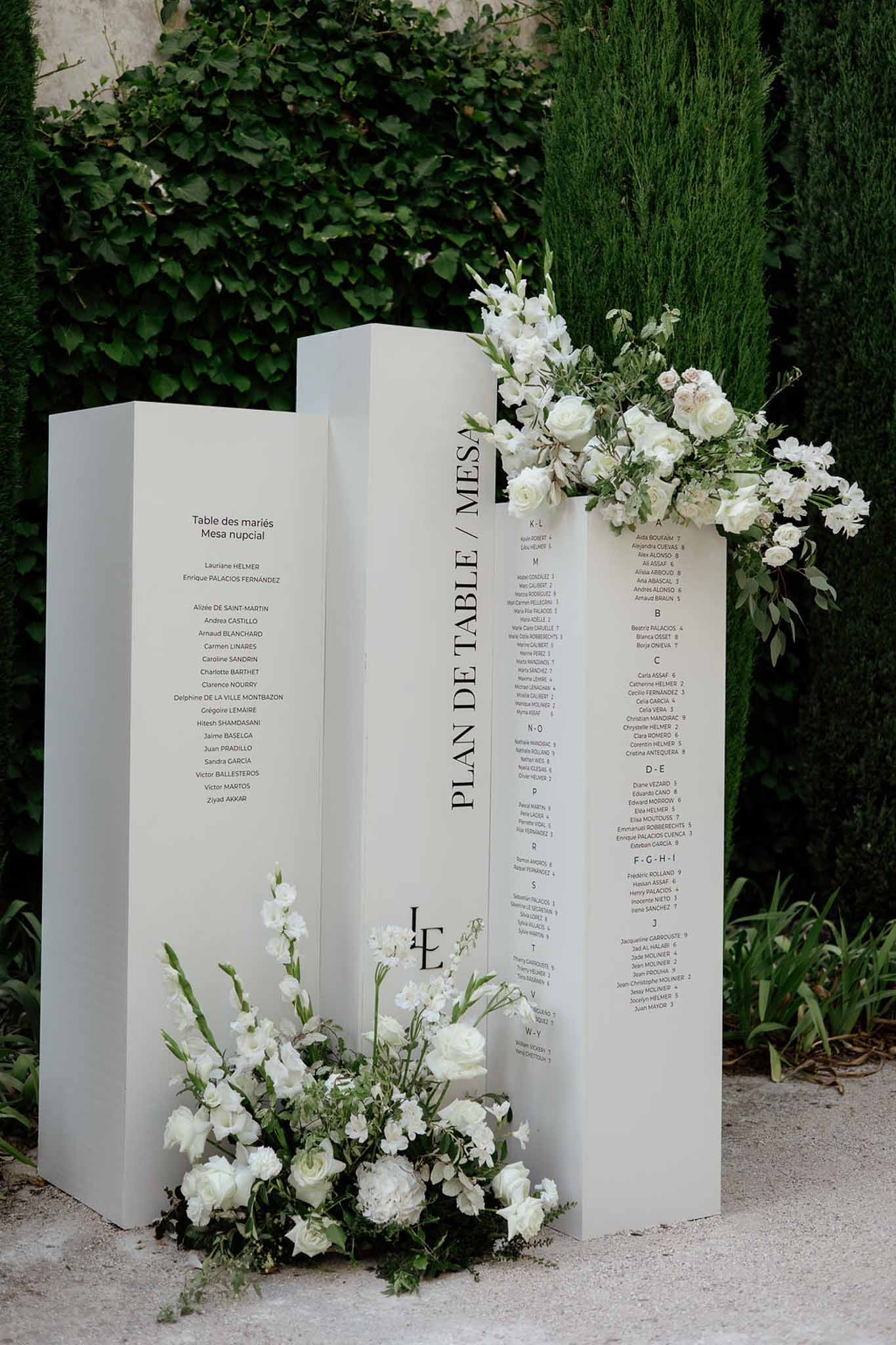 Three white column seating chart panels with serif typography and cascading white rose arrangements on gravel courtyard