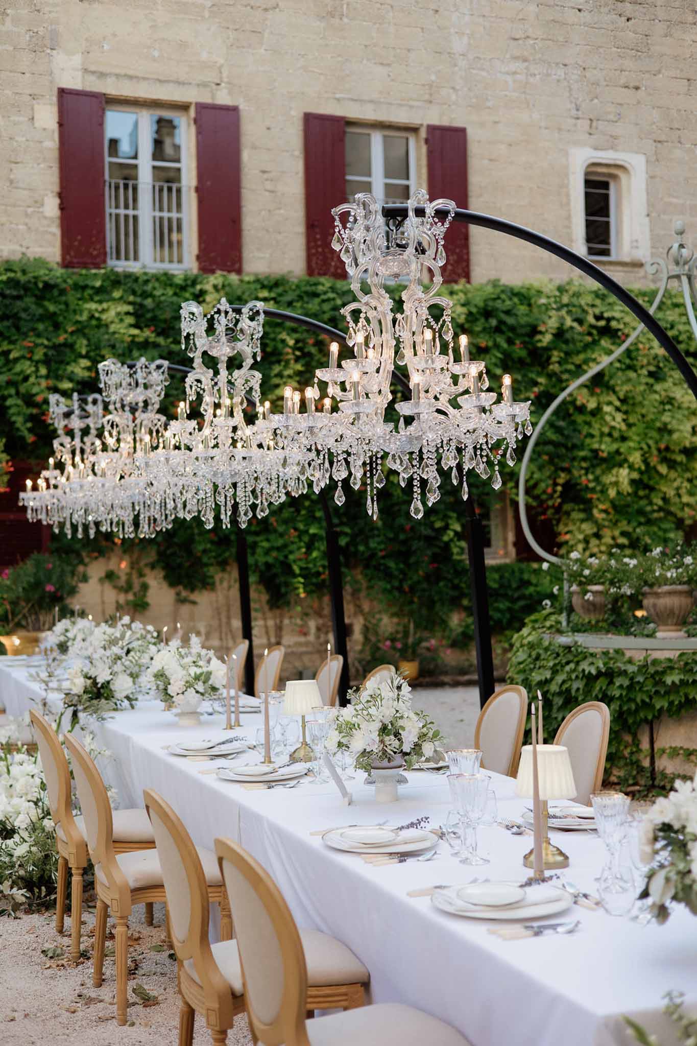 Long reception table with crystal chandeliers, white floral arrangements, and gold candle holders in a chateau courtyard