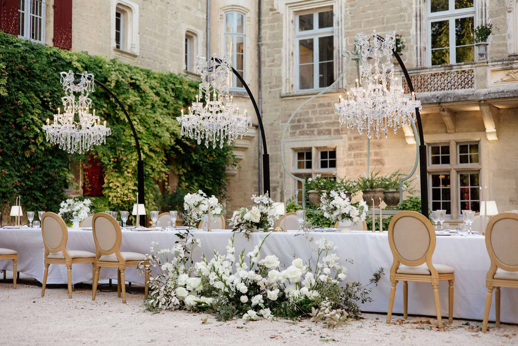Chateau courtyard reception with crystal chandeliers on arch stands, Louis XVI chairs, and white rose arrangements