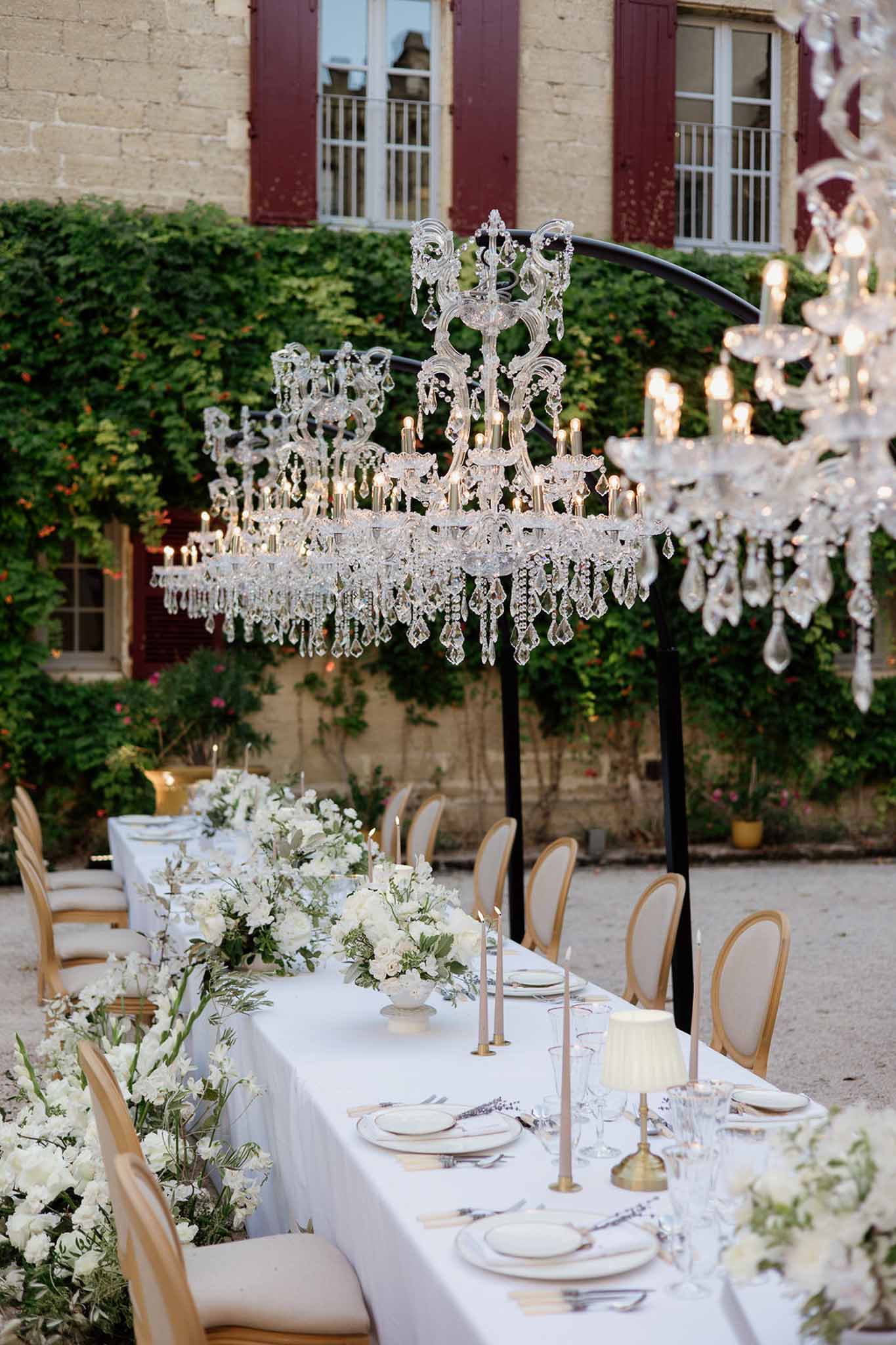 Long reception table with white roses, gold chargers, crystal chandeliers on arched stands in ivy-covered chateau courtyard