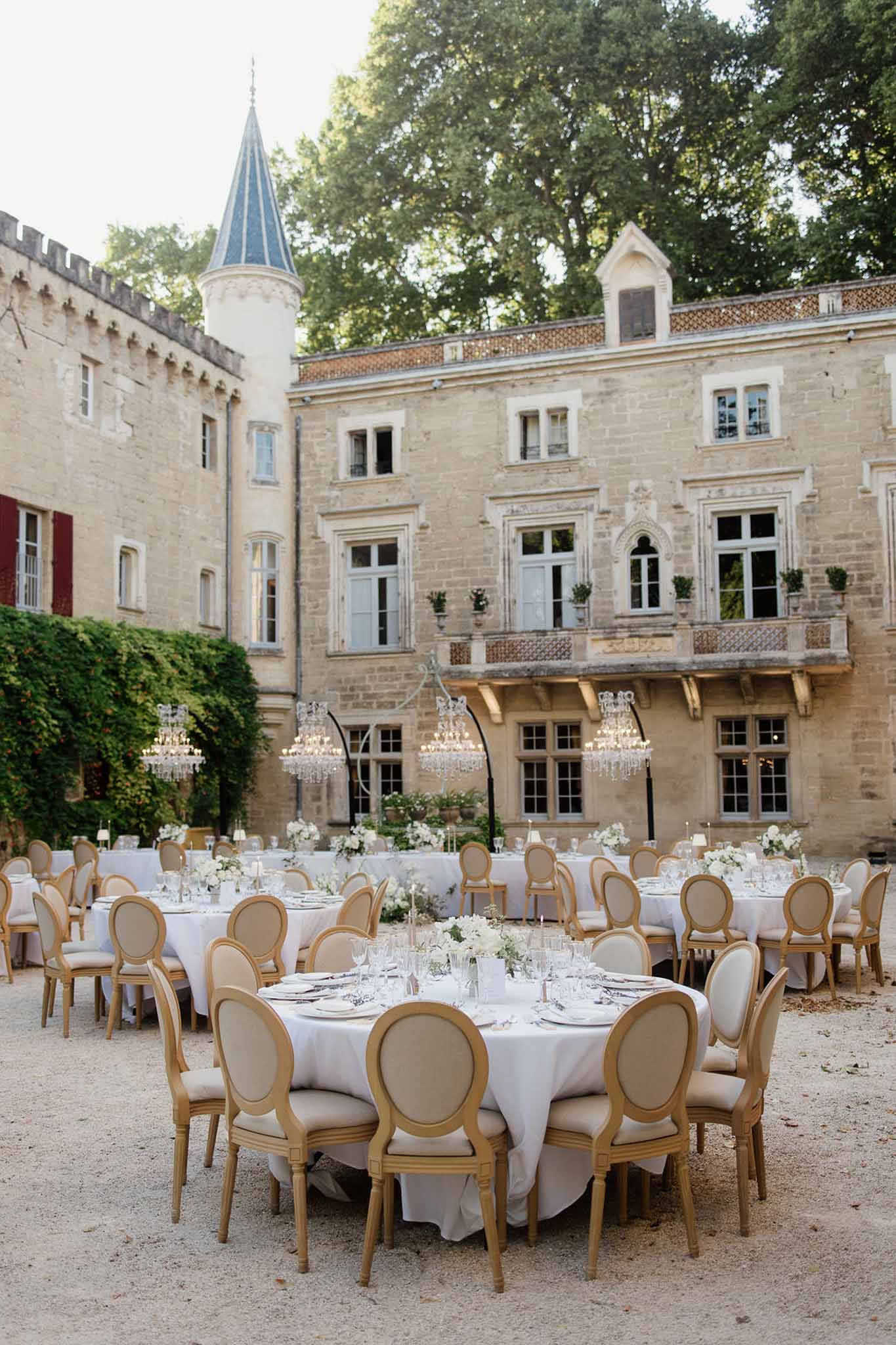 Outdoor chateau courtyard reception with round white-linen tables, gold Louis XVI chairs, and suspended crystal chandeliers