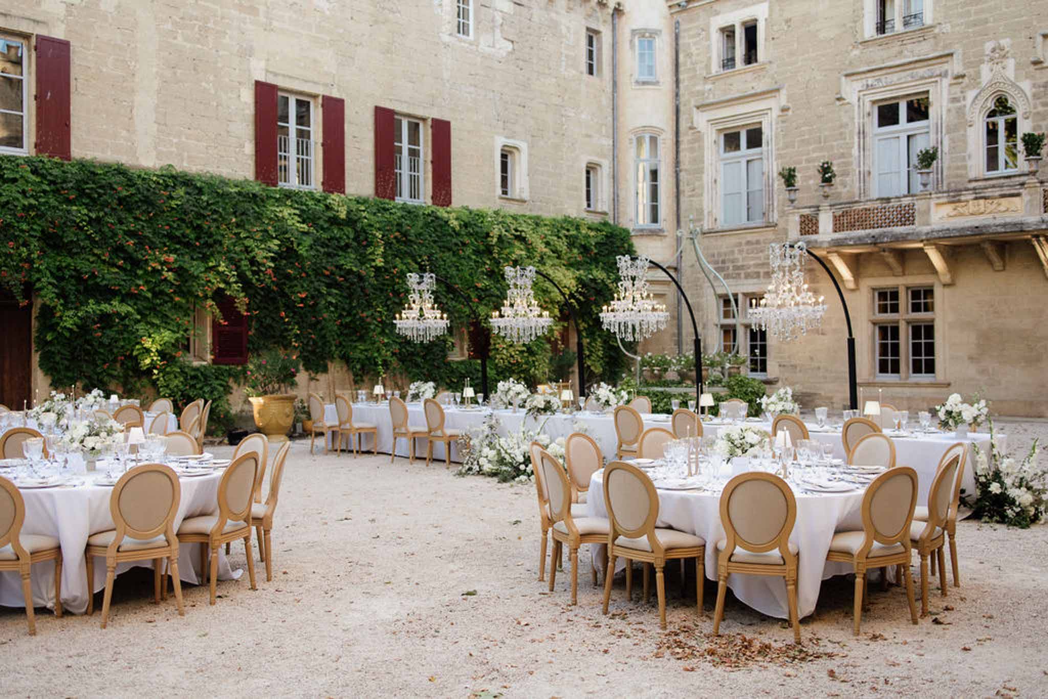 Chateau courtyard reception with round tables in white linens, gold chairs, crystal chandeliers on black stands, and white...