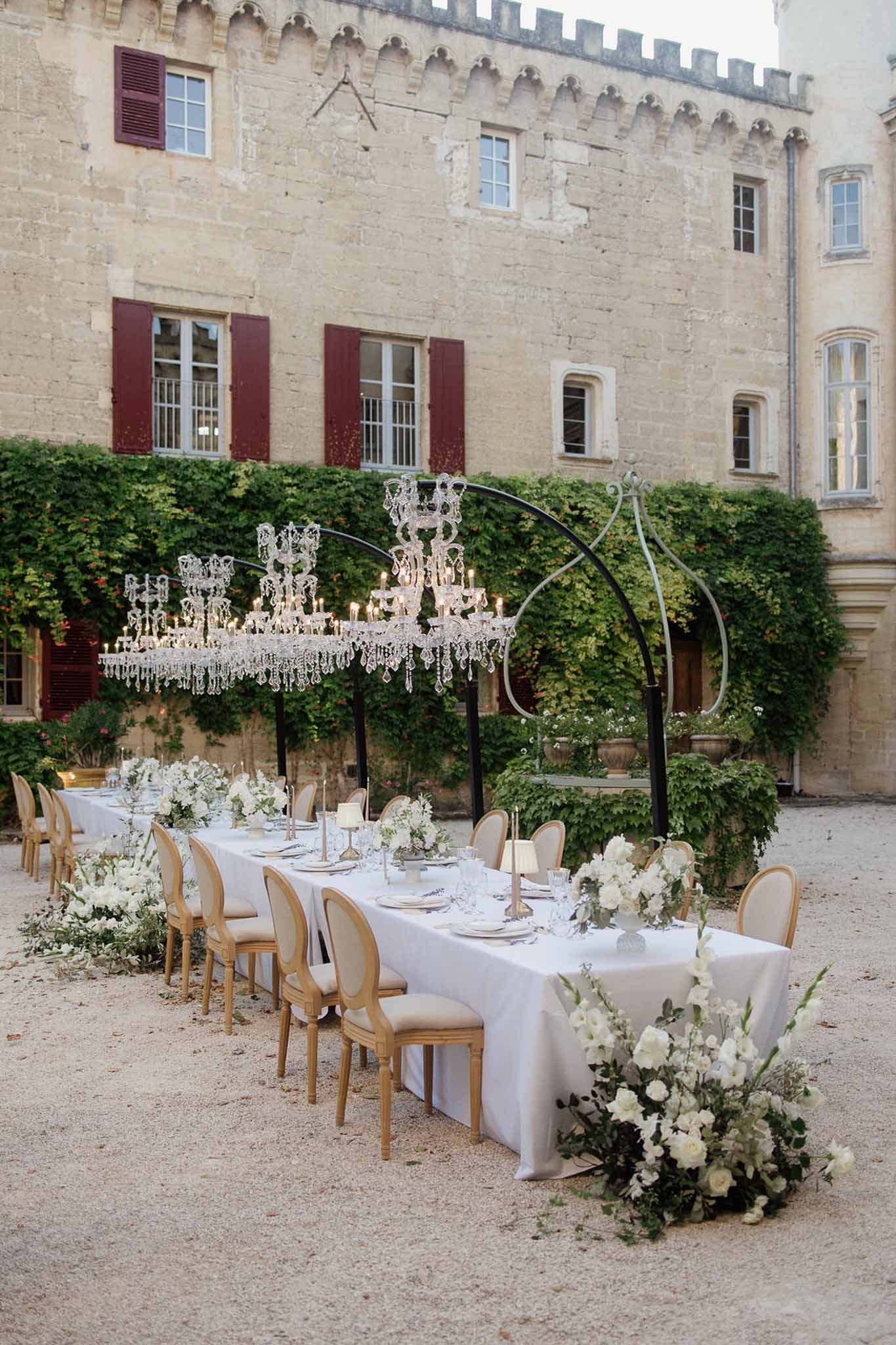 Crystal chandeliers on iron arches over white rose table with Louis XVI chairs at crenellated chateau