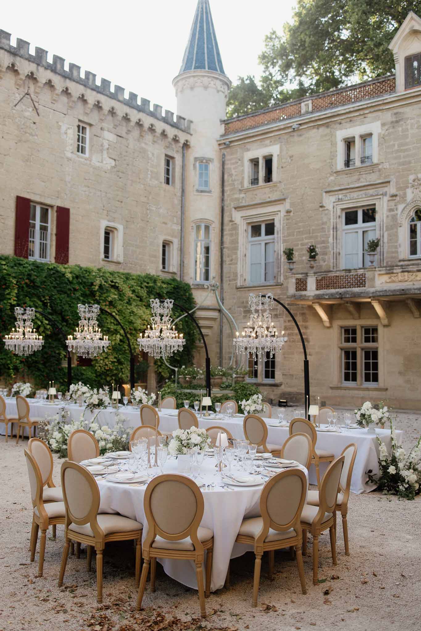 Chateau courtyard reception with round white-linen tables, gold chairs, and crystal chandeliers on stands