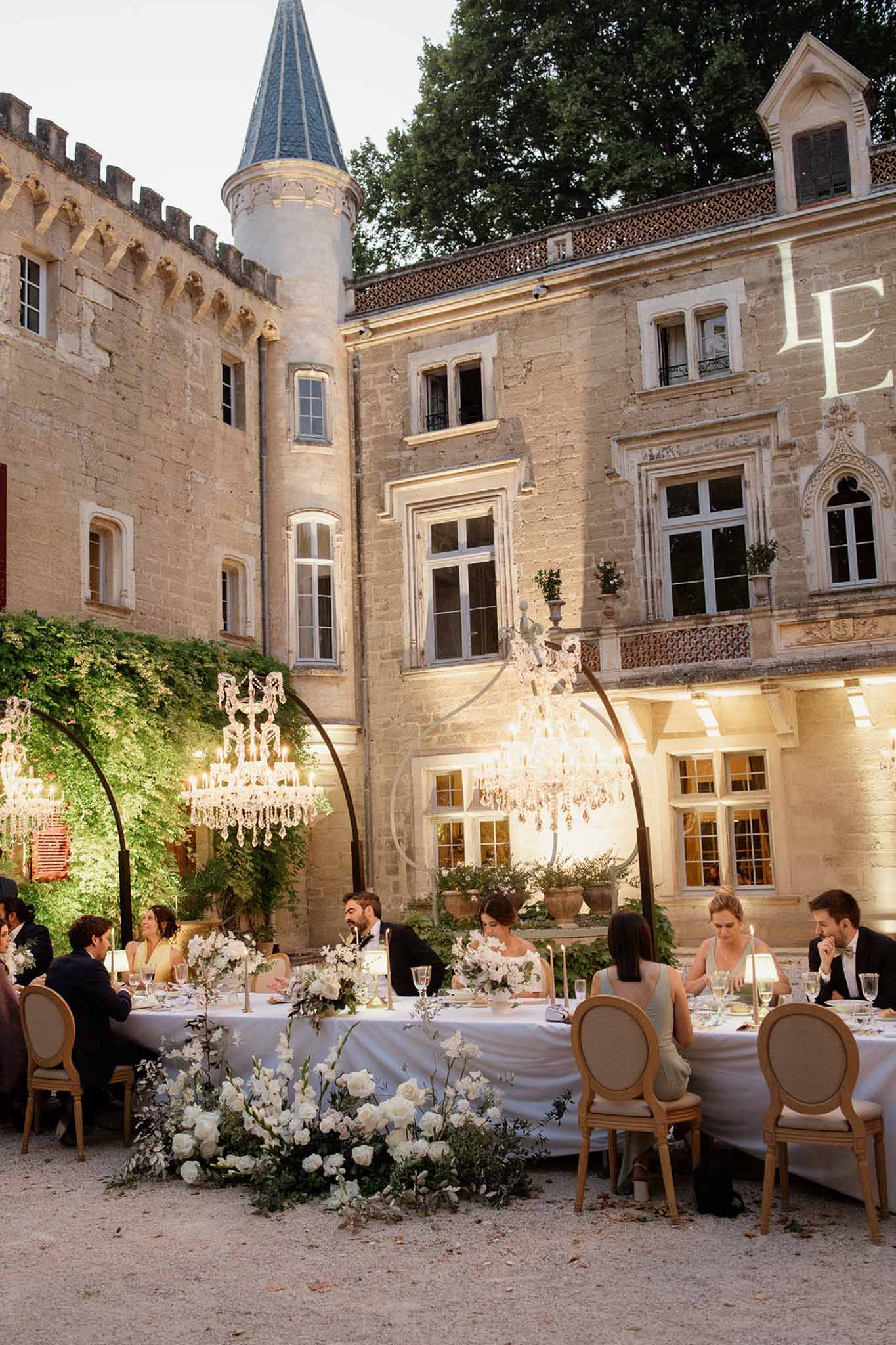 Crystal chandeliers on metal arches over long table with white roses at turreted chateau at dusk