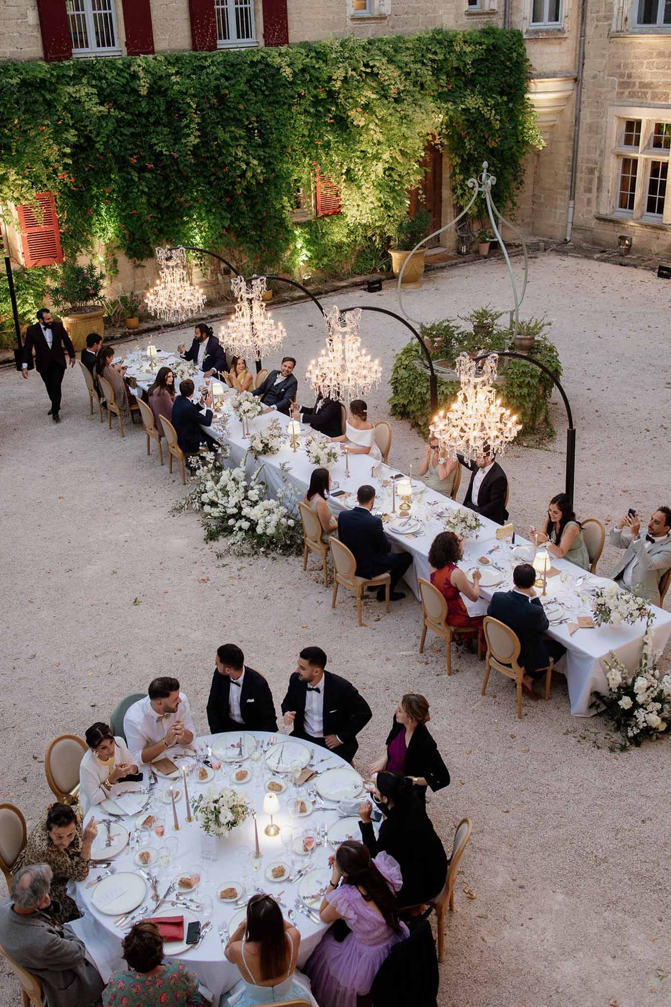 Elevated view of reception with crystal chandeliers on iron arches white floral runner gold chairs and forty guests