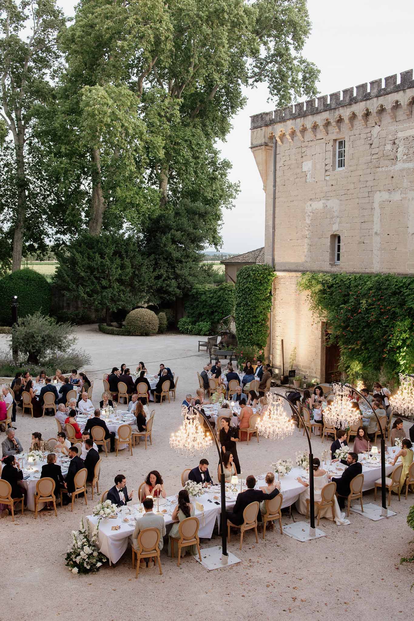 Elevated view of outdoor chateau courtyard reception with round tables, candelabra arc stands, and seated guests at dusk