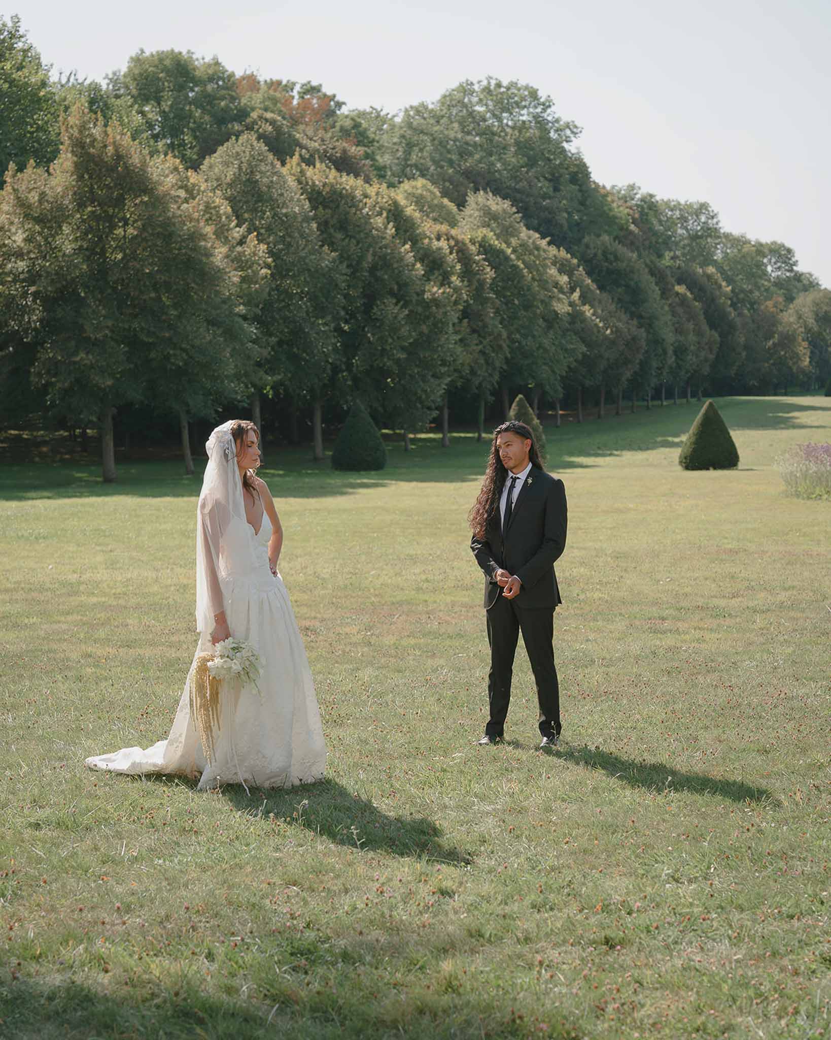 Bride and groom standing apart on a chateau lawn with trimmed topiary and mature trees