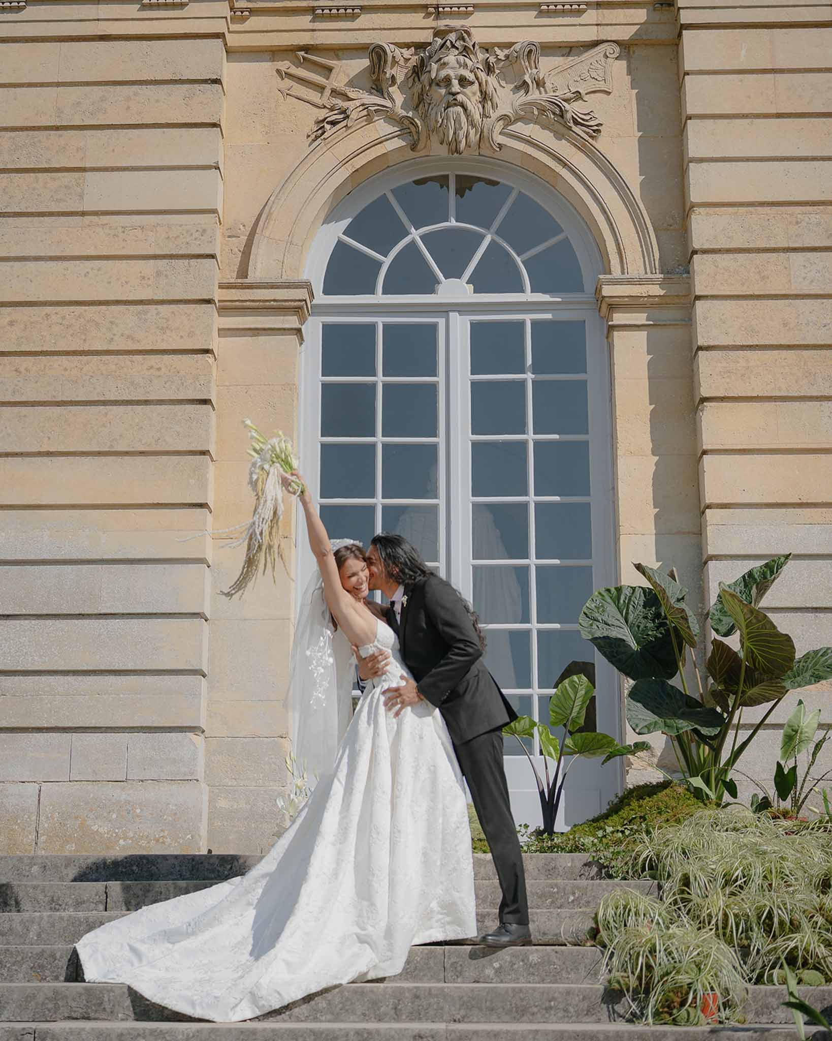 Groom dipping bride as she raises bouquet overhead on limestone chateau steps with carved mascaron archway