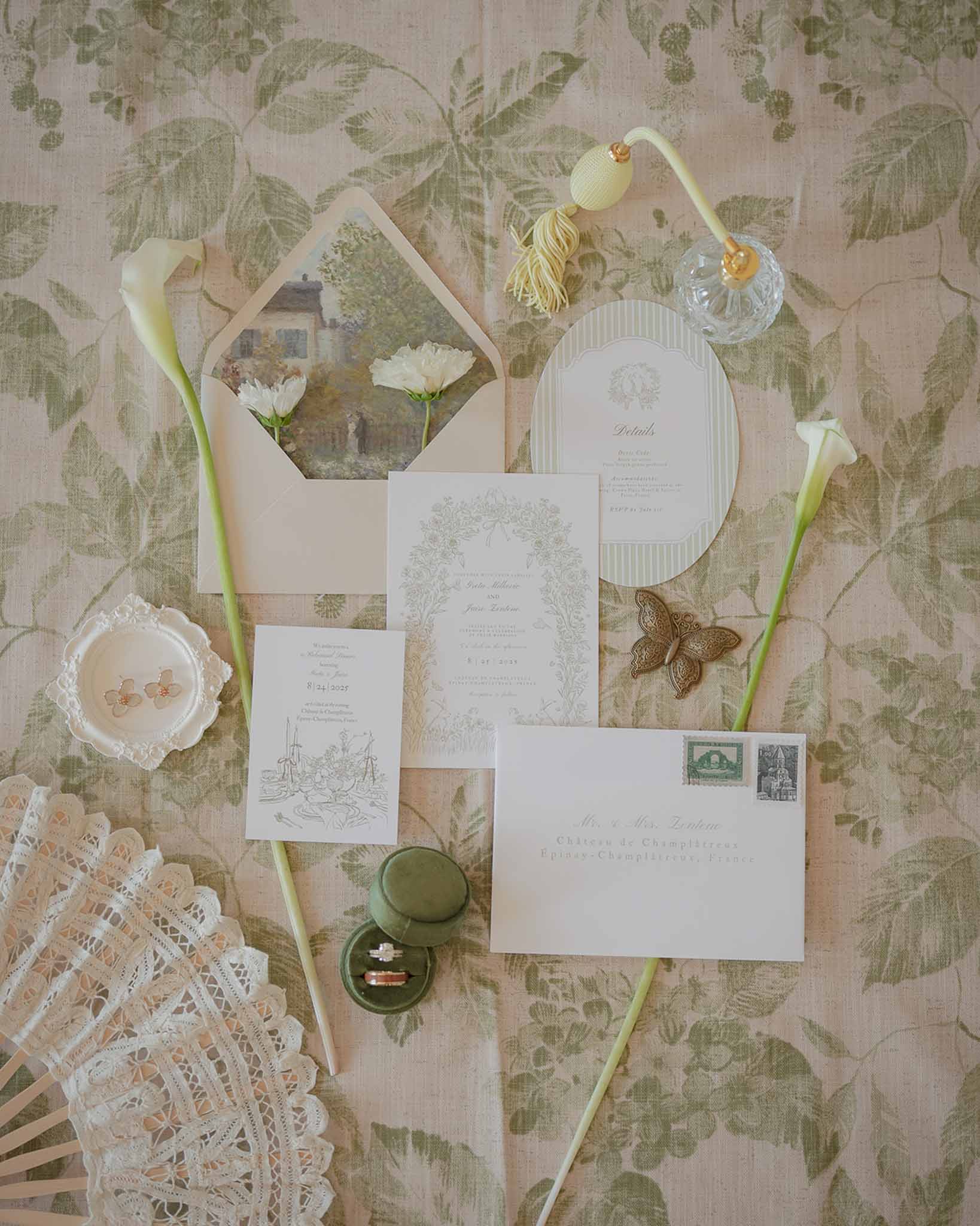 Flat lay of sage botanical invitation suite with velvet ring box, crystal perfume, and calla lilies