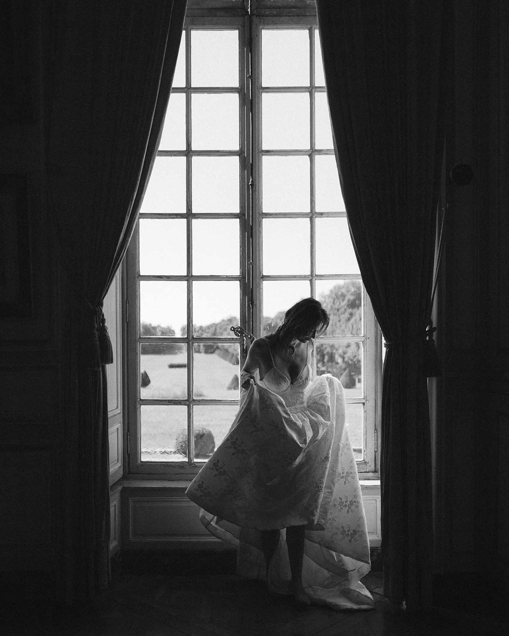 Black and white silhouette of bride adjusting embroidered gown before tall French windows with garden view beyond