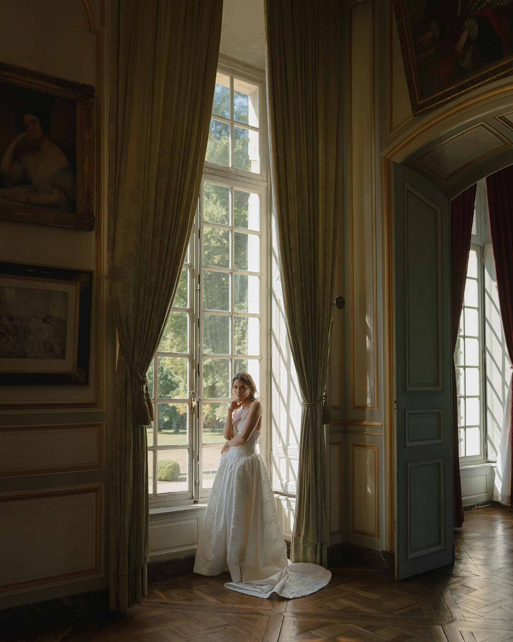 Bride in white ball gown silhouetted against tall French windows inside a chateau with sage green drapes