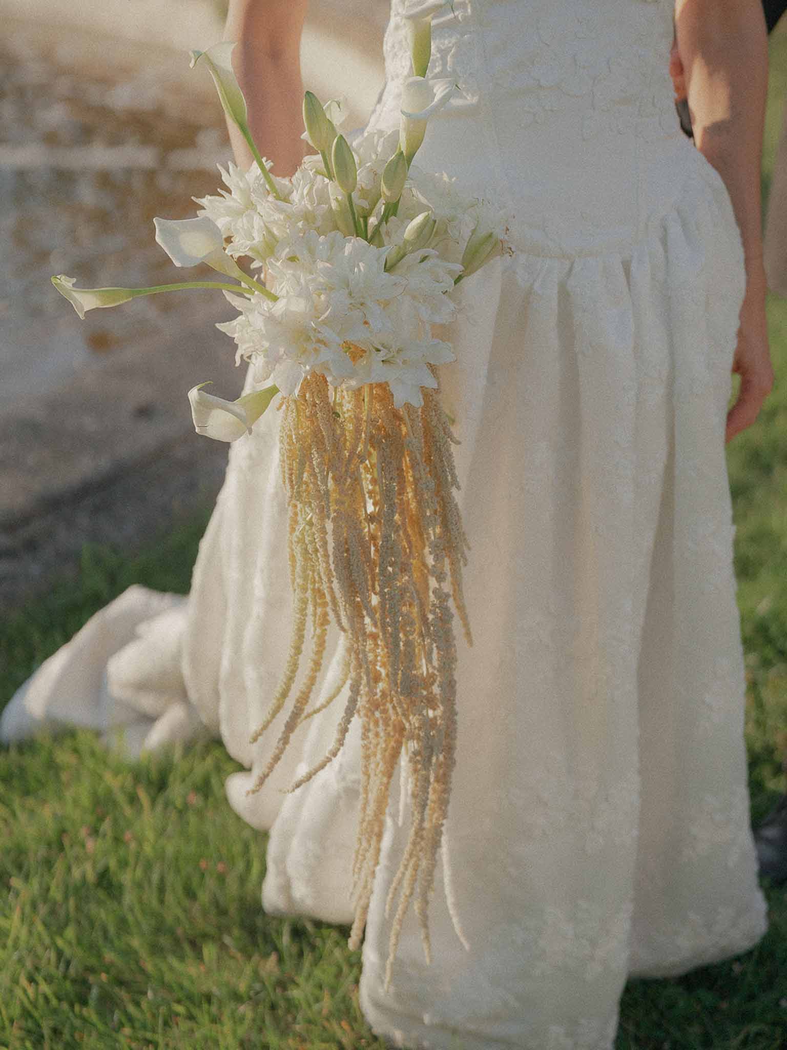 Cascading bouquet of white calla lilies and tuberose with golden amaranthus trails held against textured gown at sunset