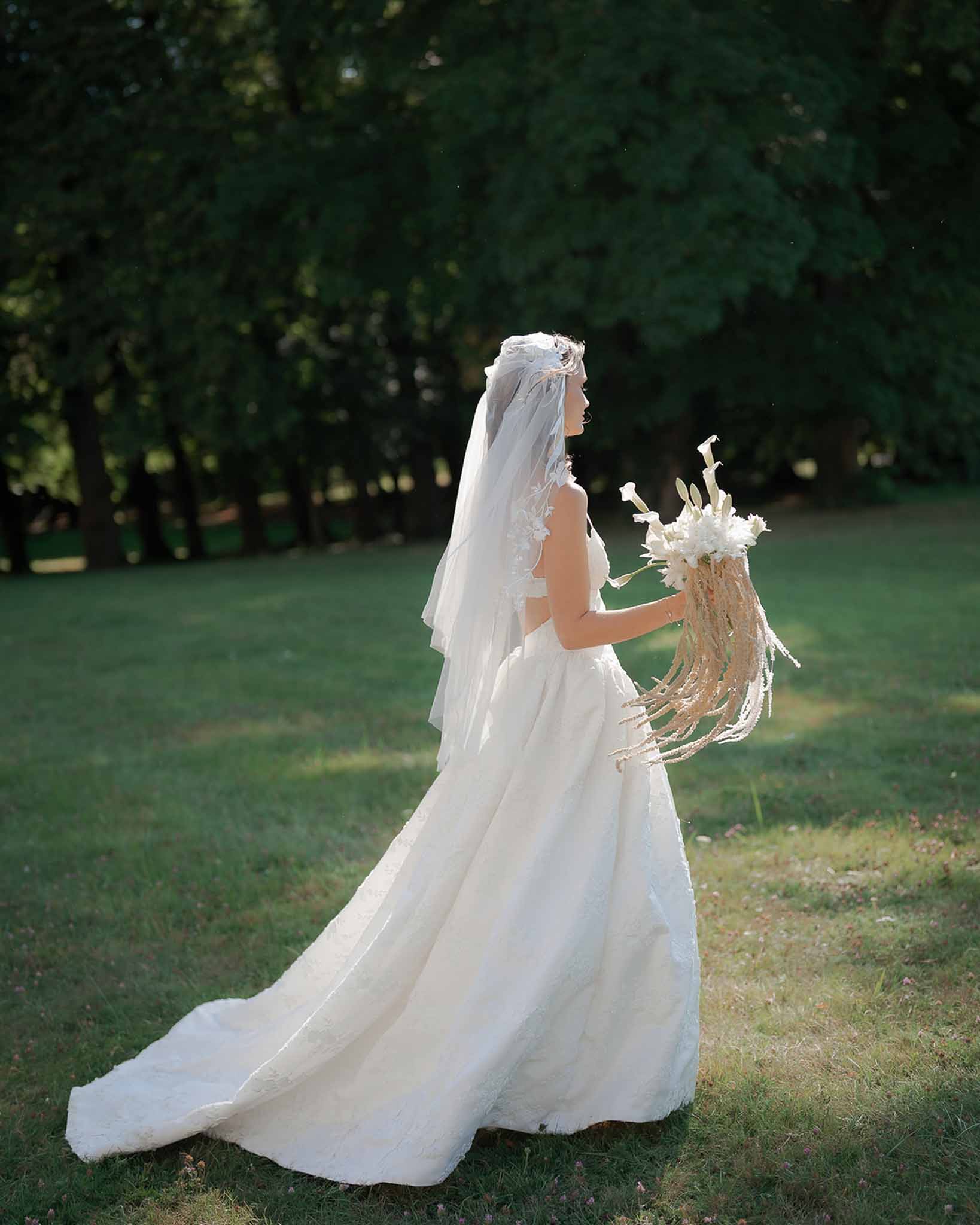 Bride in ivory textured ballgown with floral-applique veil holding cascading calla lily and dried stem bouquet in garden
