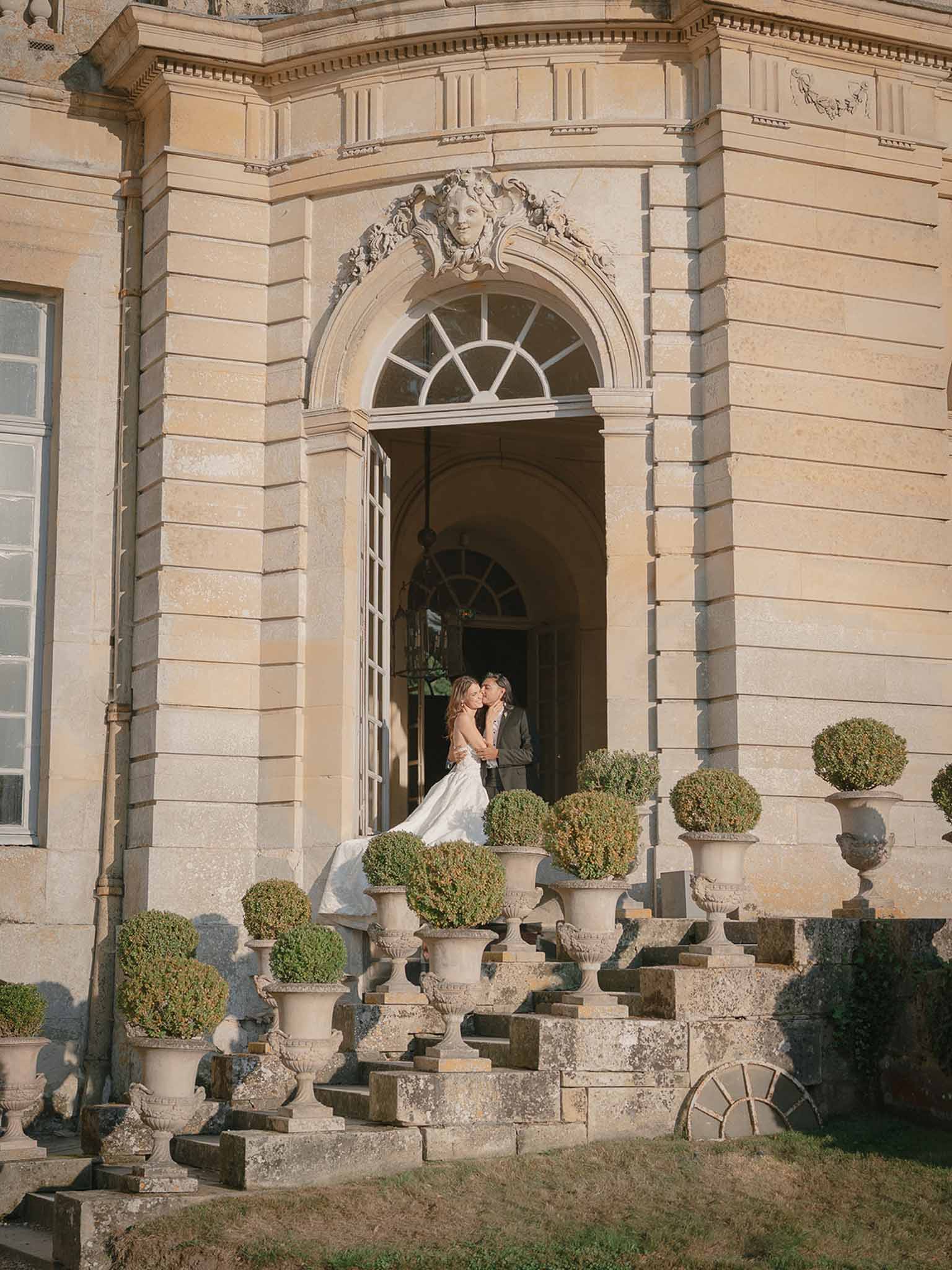 Bride and groom kiss in arched doorway of golden limestone chateau with topiary-lined stone staircase