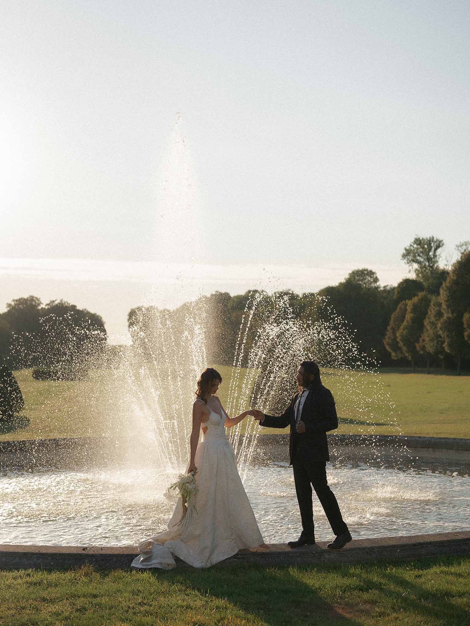 Bride and groom holding hands beside an ornamental fountain backlit by golden hour sunlight in estate gardens