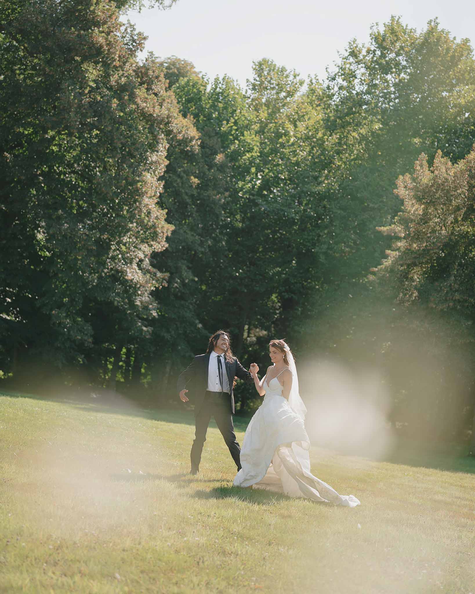 Couple running and laughing on lawn with cathedral veil and golden lens flare at sunset