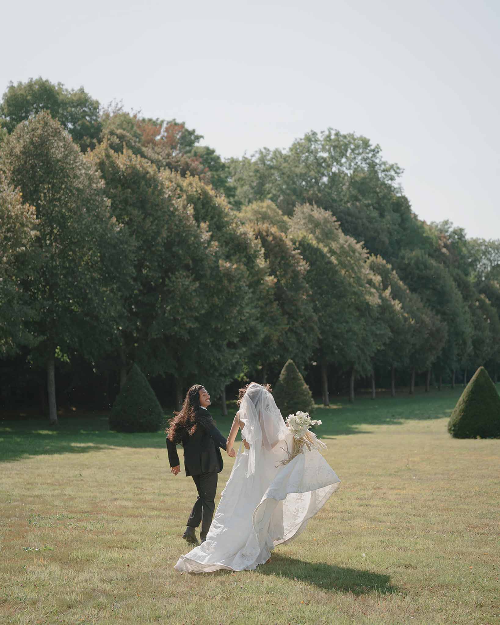 Couple running on chateau lawn brides cathedral veil billowing groom laughing with conical topiary and tree-lined allee