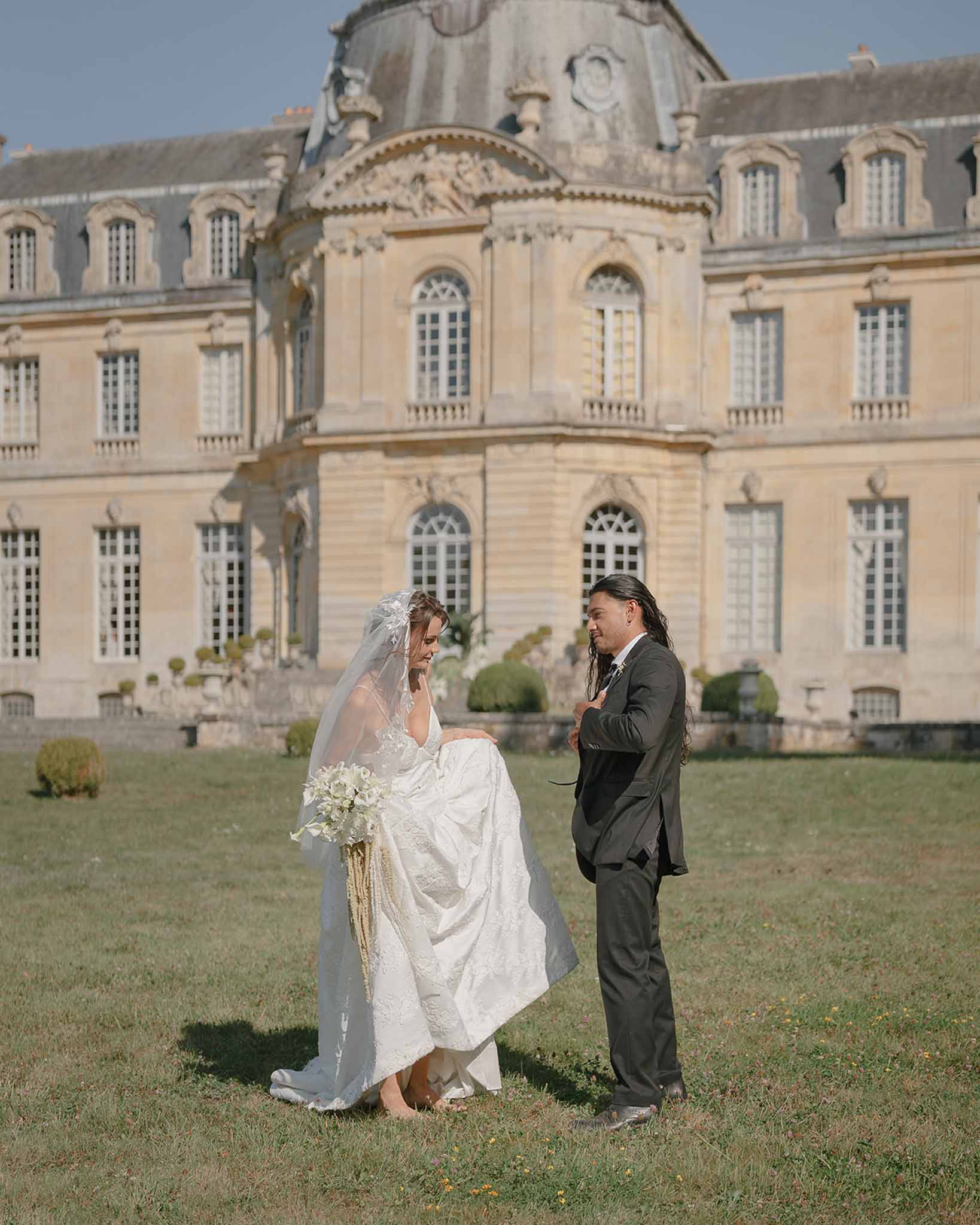 Bride in ivory lace gown with cathedral veil and groom standing on lawn before domed French chateau
