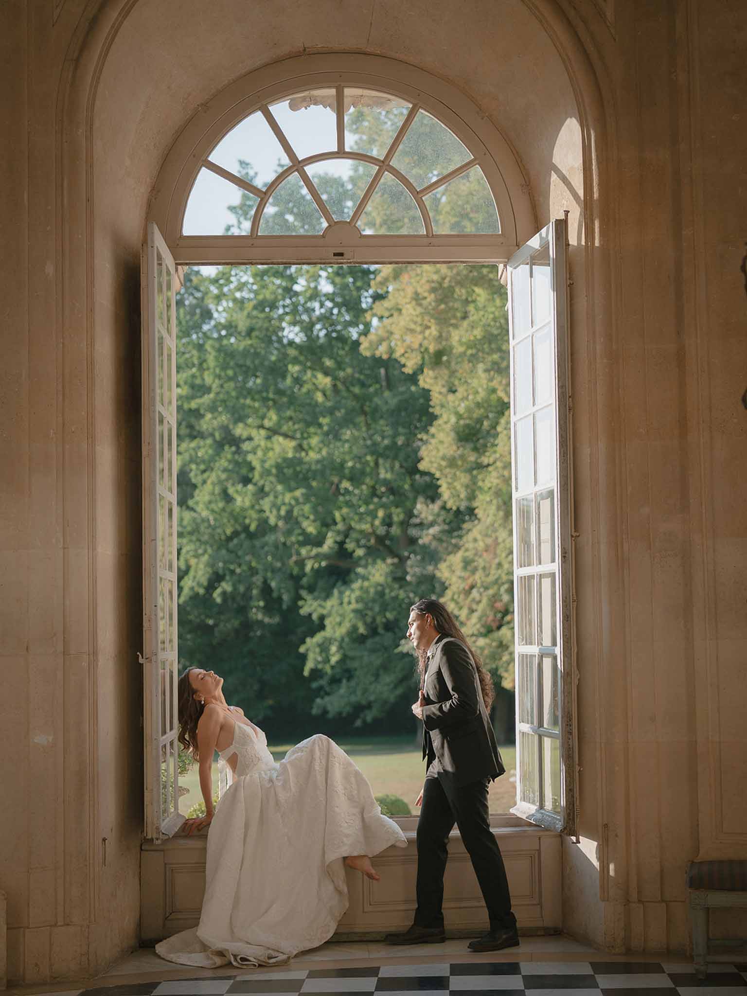 Bride seated barefoot in ball gown with groom standing by arched French door in chateau