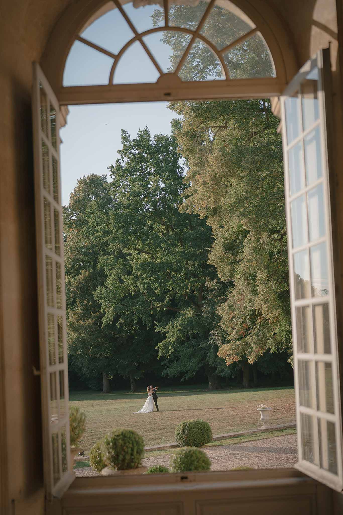 Couple framed through tall French doors with fanlight arch embracing on distant lawn in golden afternoon light