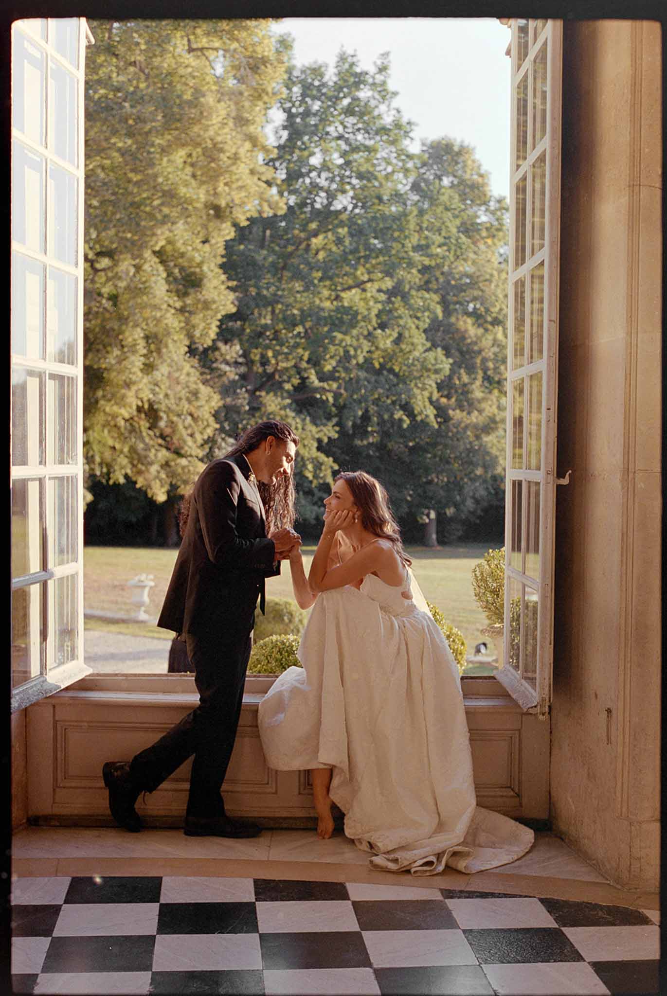 Groom kissing bride's hand as she sits on window ledge inside chateau with checkered marble floor