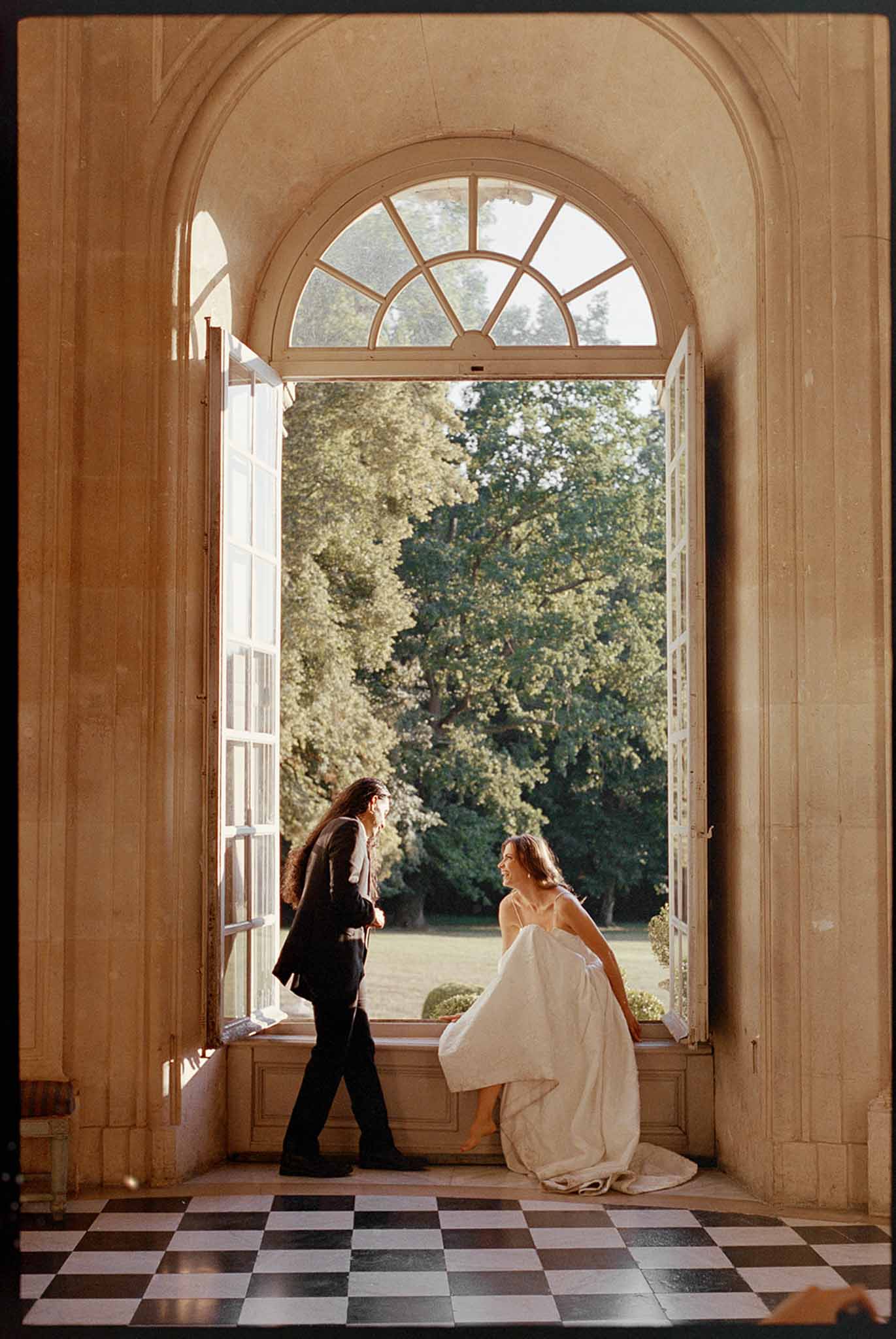 Film-toned portrait of bride on window sill chatting with groom framed by stone arch at chateau