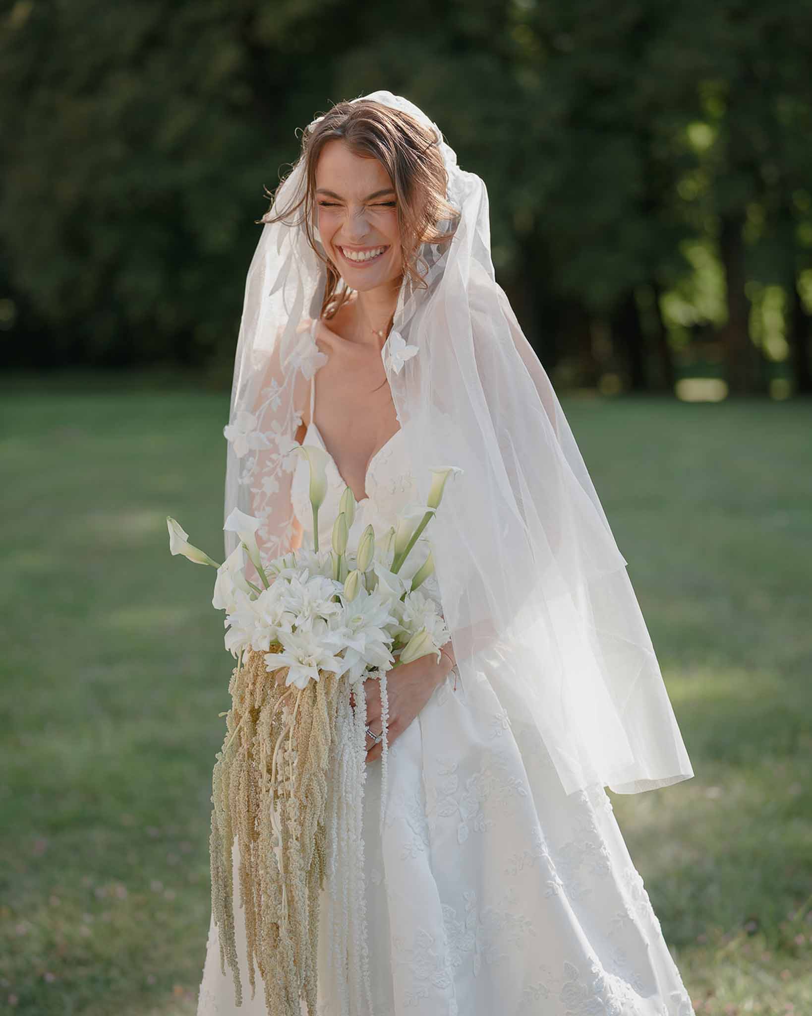 Bride laughing in lace gown and embellished veil holding cascading lily and dried amaranthus bouquet