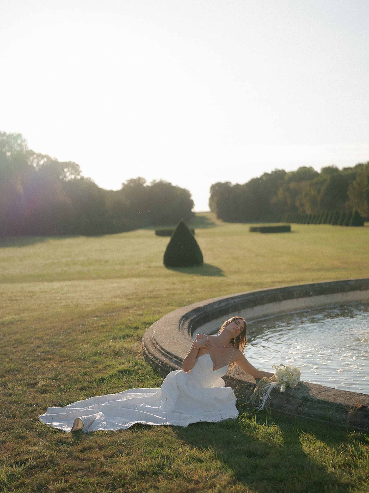 Bride in fitted spaghetti-strap gown seated at ornamental pond with topiary garden at golden hour