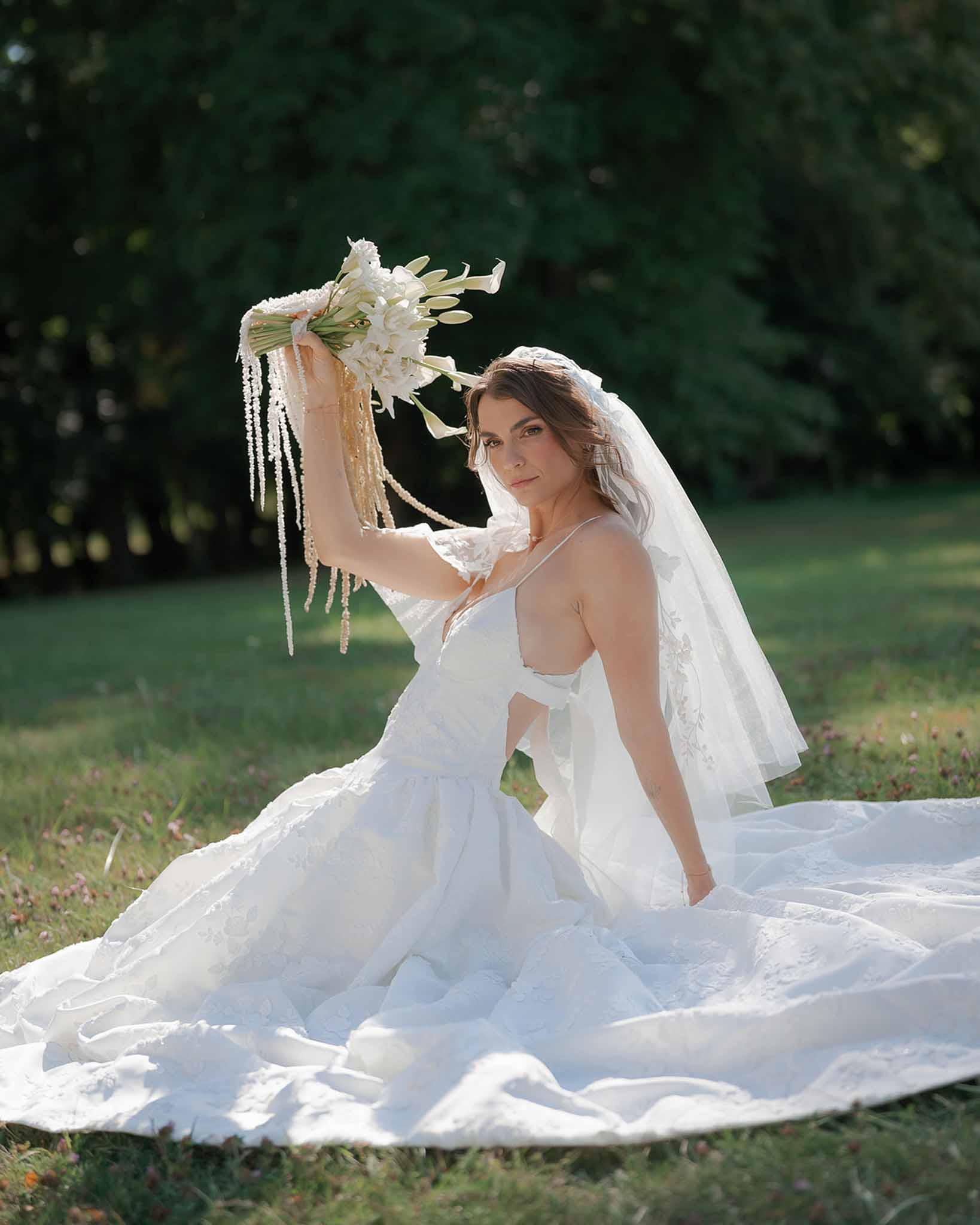 Bride seated on grass in ivory floral applique gown holding a calla lily bouquet with trailing pearl strands above her head