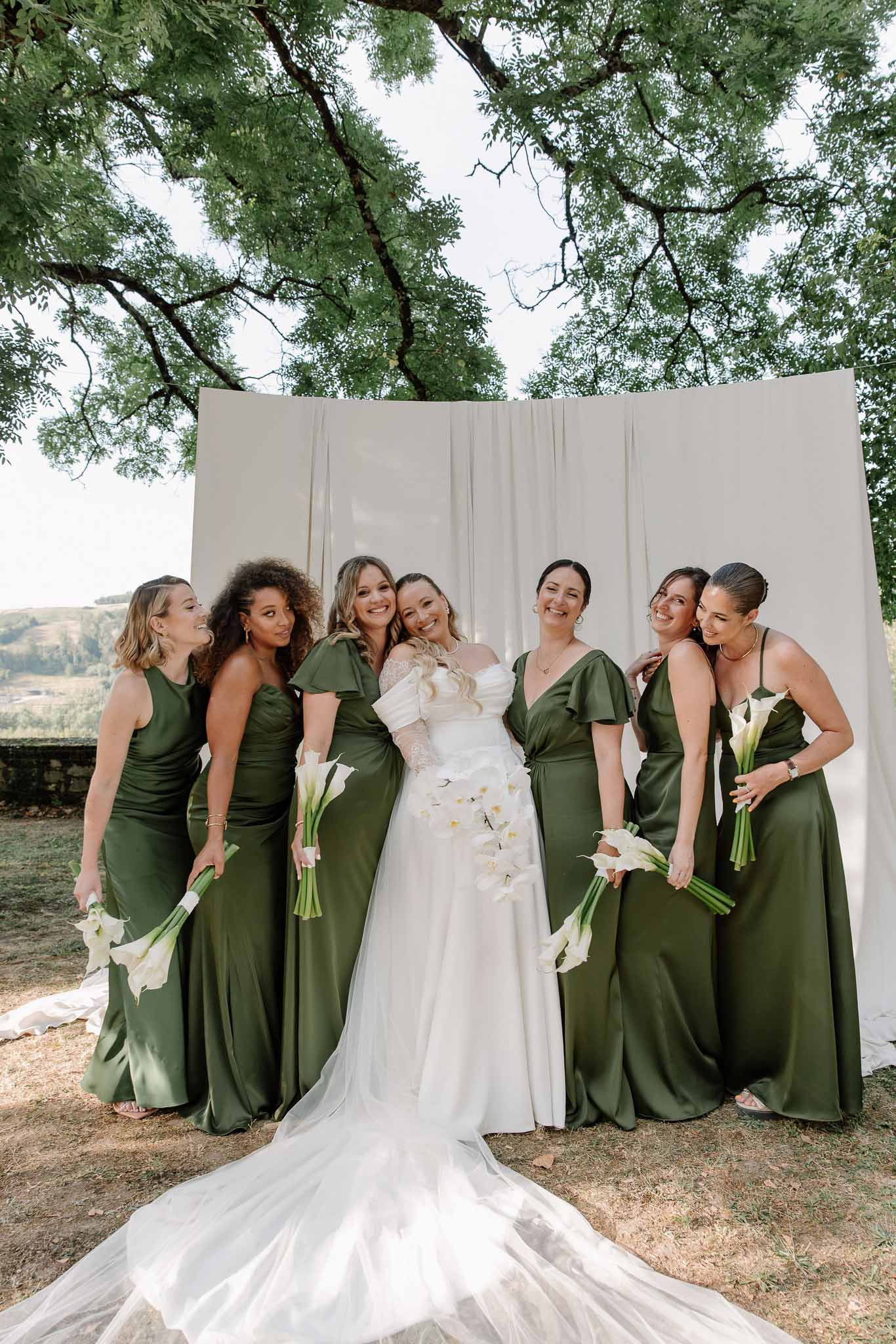 Bride with six bridesmaids in olive green satin dresses laughing together outdoors