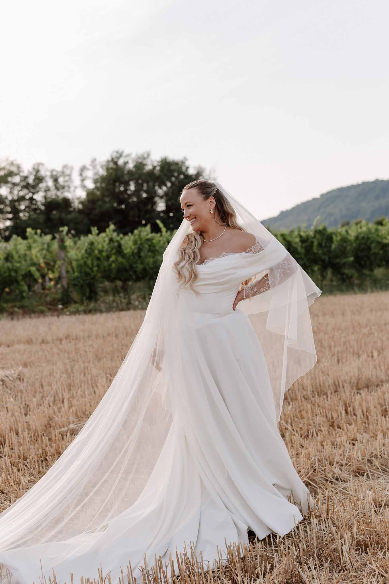 Bride in ivory A-line gown with cathedral-length lace veil standing in harvested wheat field with vineyard backdrop