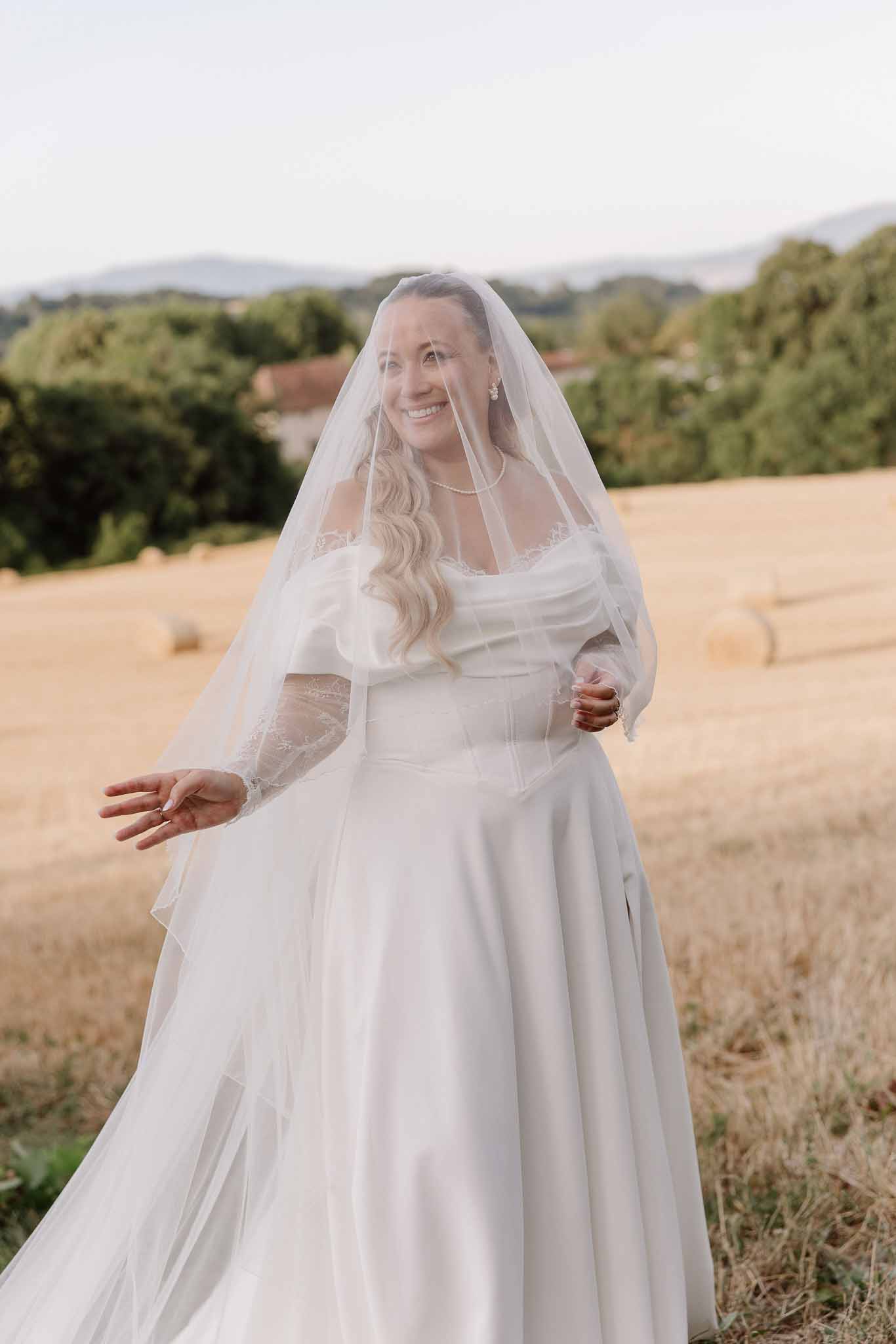 Bride in off-shoulder lace-sleeve gown with cathedral veil and pearls turning in golden-hour harvest field