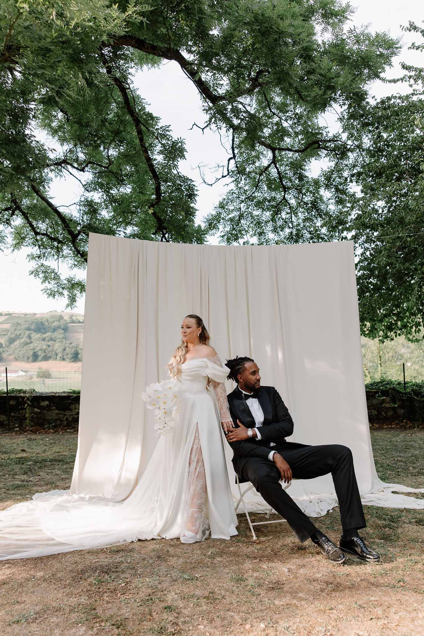 Editorial couple portrait against ivory drape outdoors bride in puff-sleeve gown with orchid bouquet groom seated in tuxedo