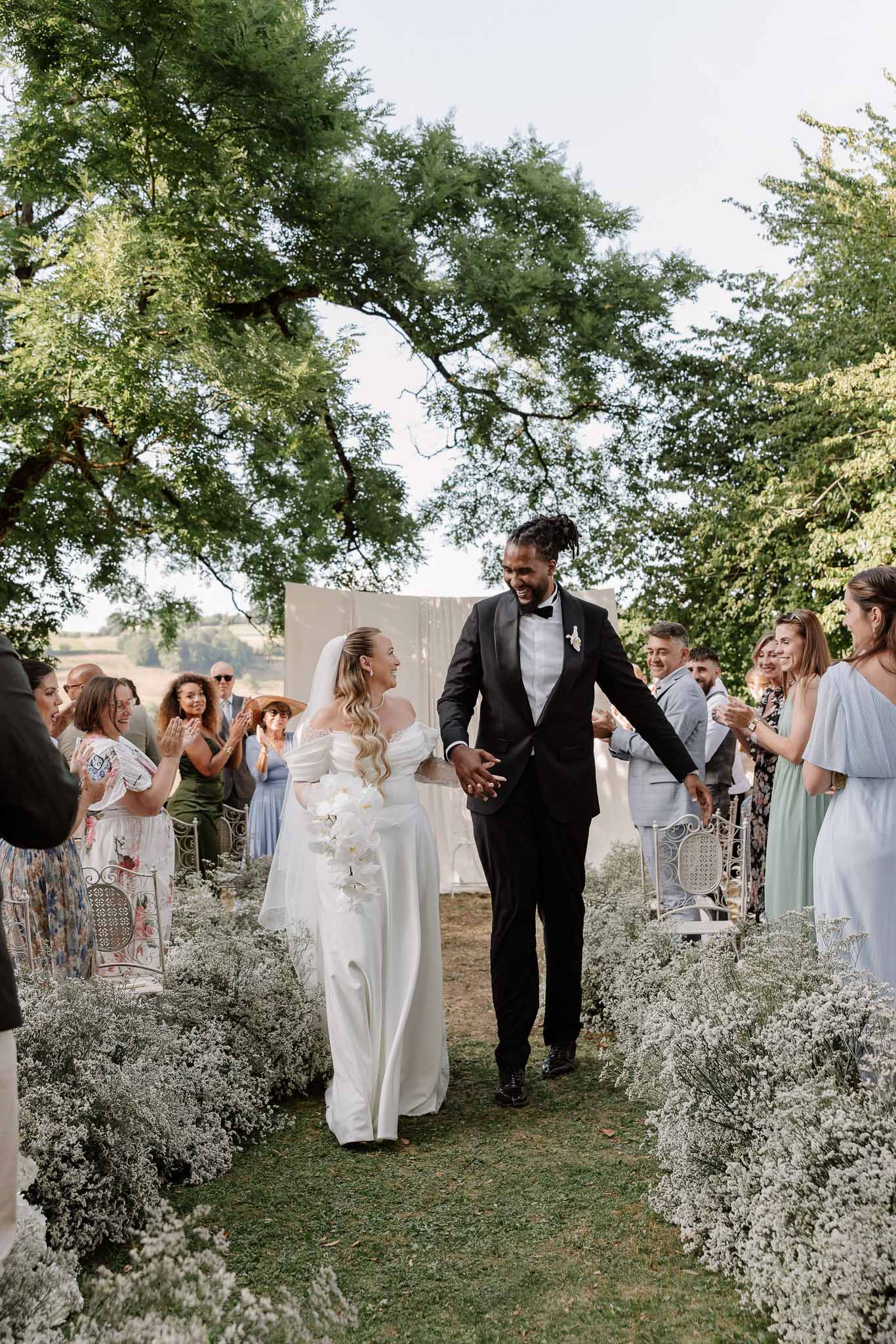 Couple walks up aisle past baby's breath borders as guests applaud from white metal chairs on lawn