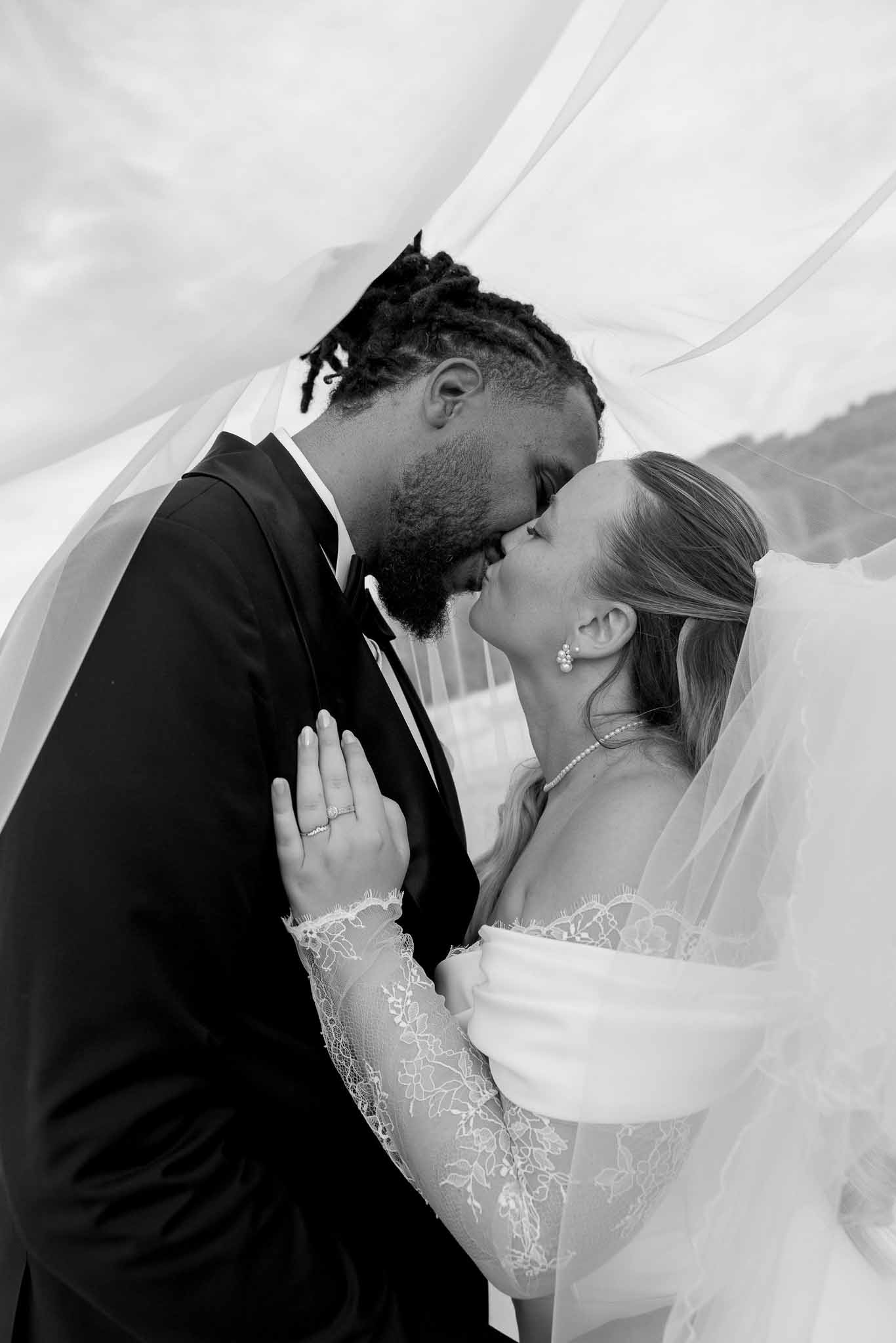 Black-and-white portrait of bride and groom kissing beneath a billowing veil with lace dress detail