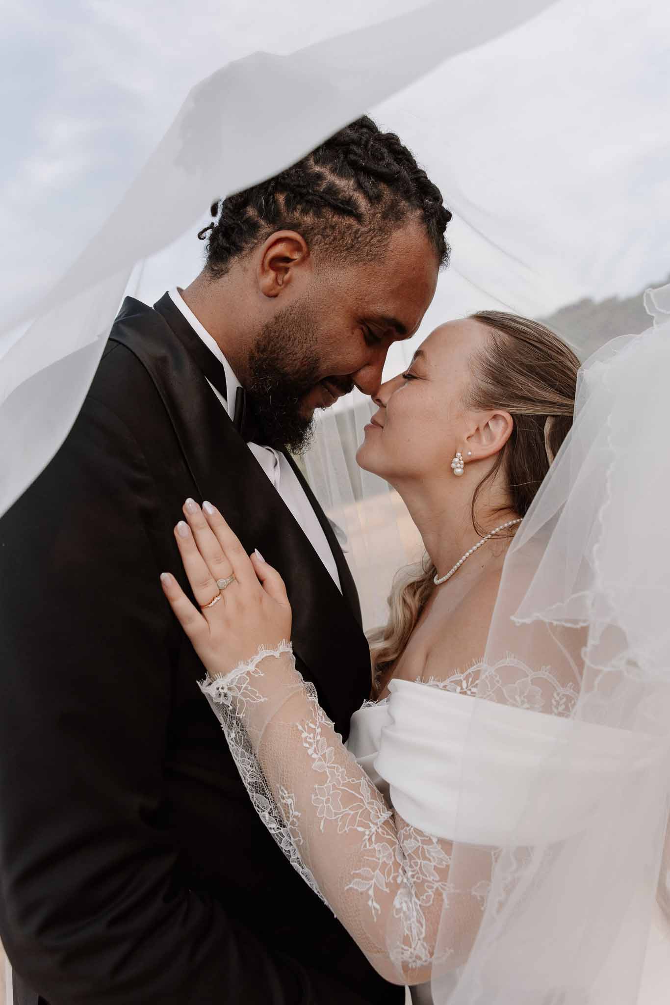 Close-up of couple touching foreheads wrapped in billowing veil, bride in lace sleeves with pearl jewelry