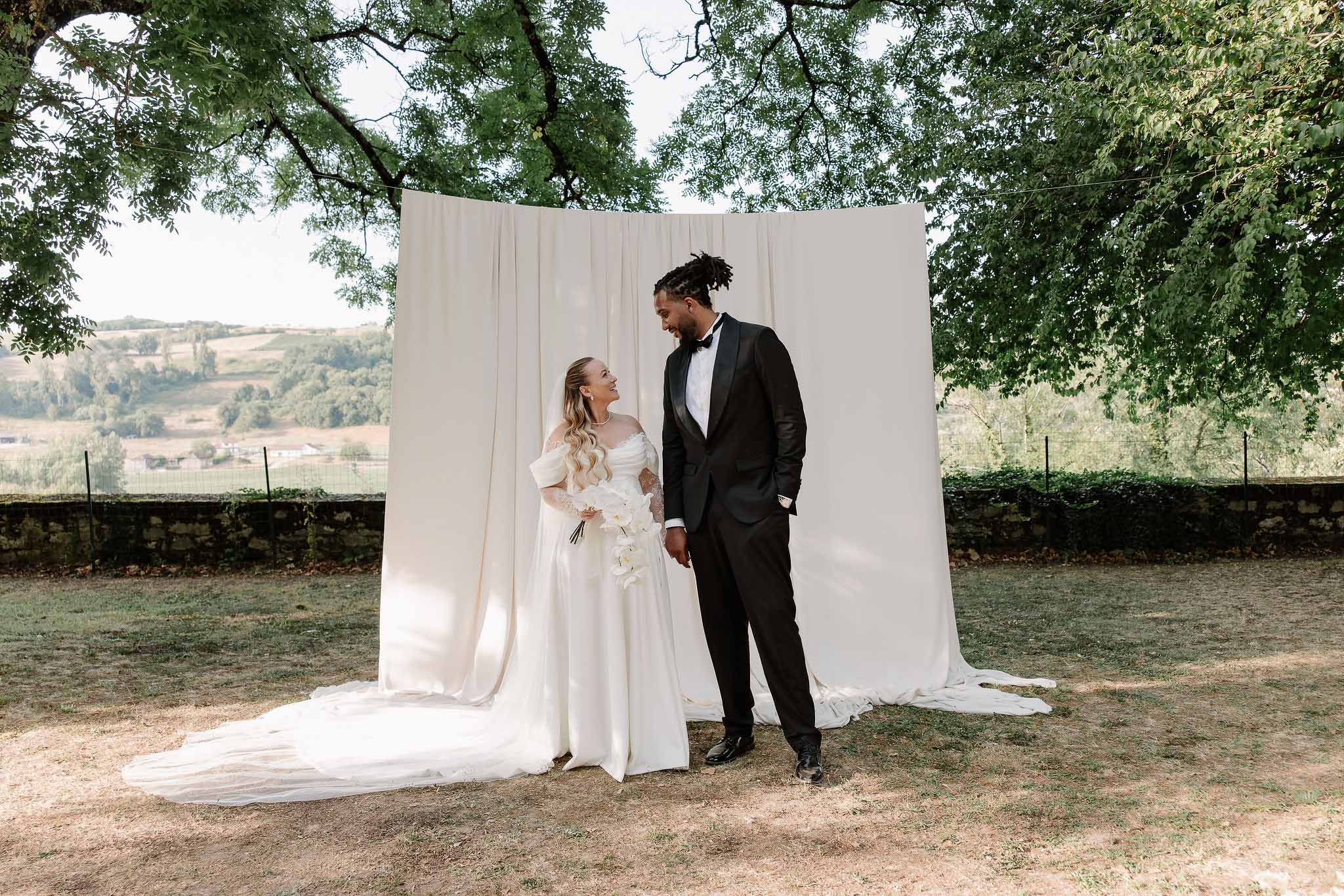 Bride in off-shoulder white satin gown and groom in black tuxedo facing each other before cream draped backdrop outdoors