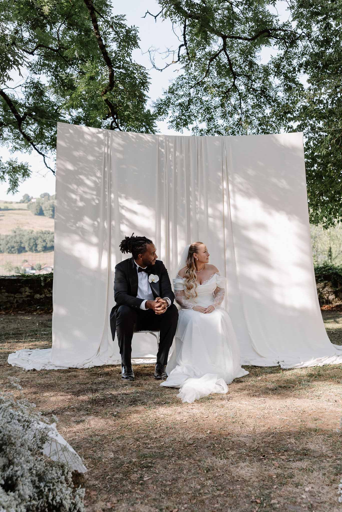 Couple seated on chairs before white draped backdrop with bride in off-shoulder lace gown and veil