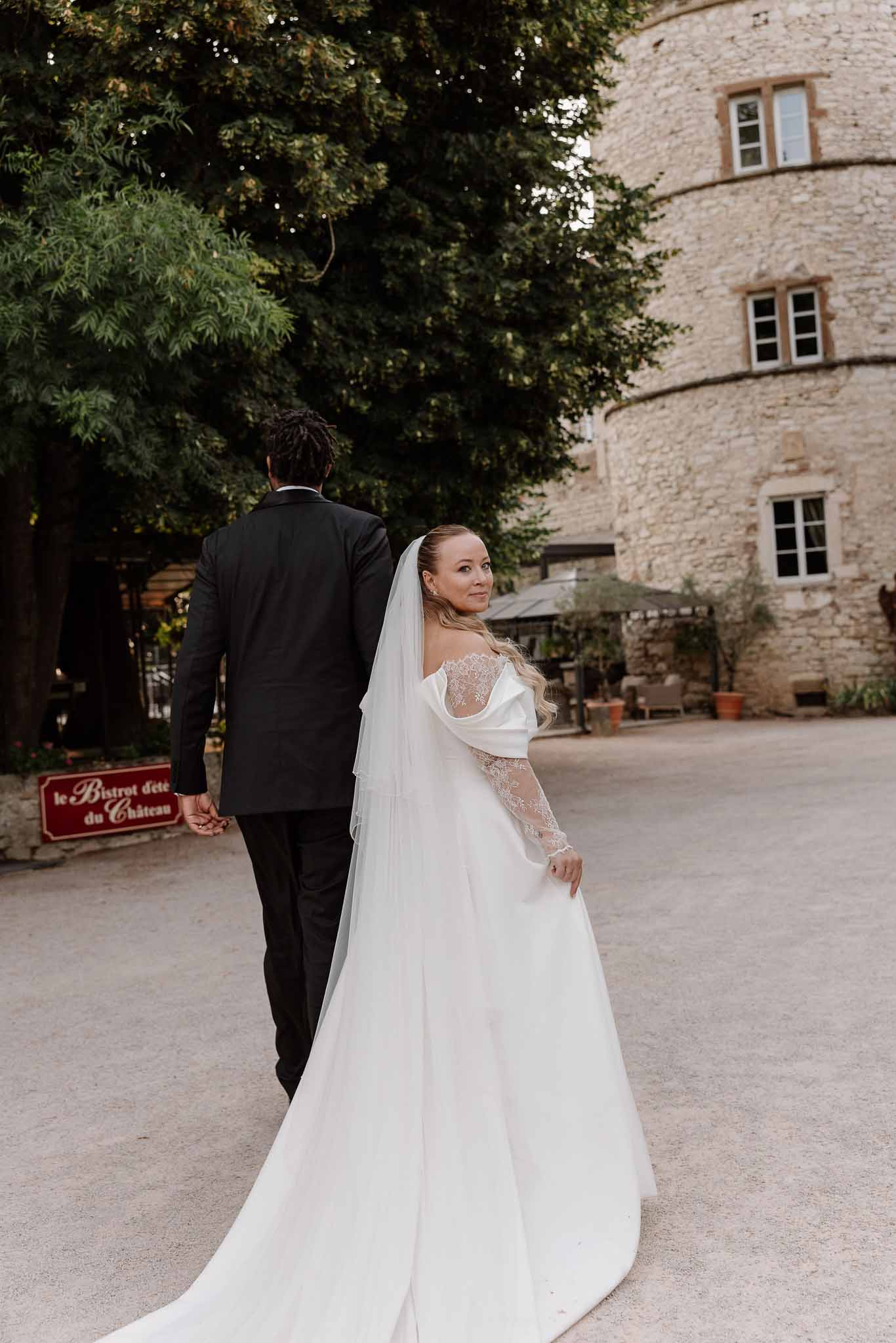 Bride in off-shoulder lace gown with veil turning back as groom leads through chateau courtyard with tower
