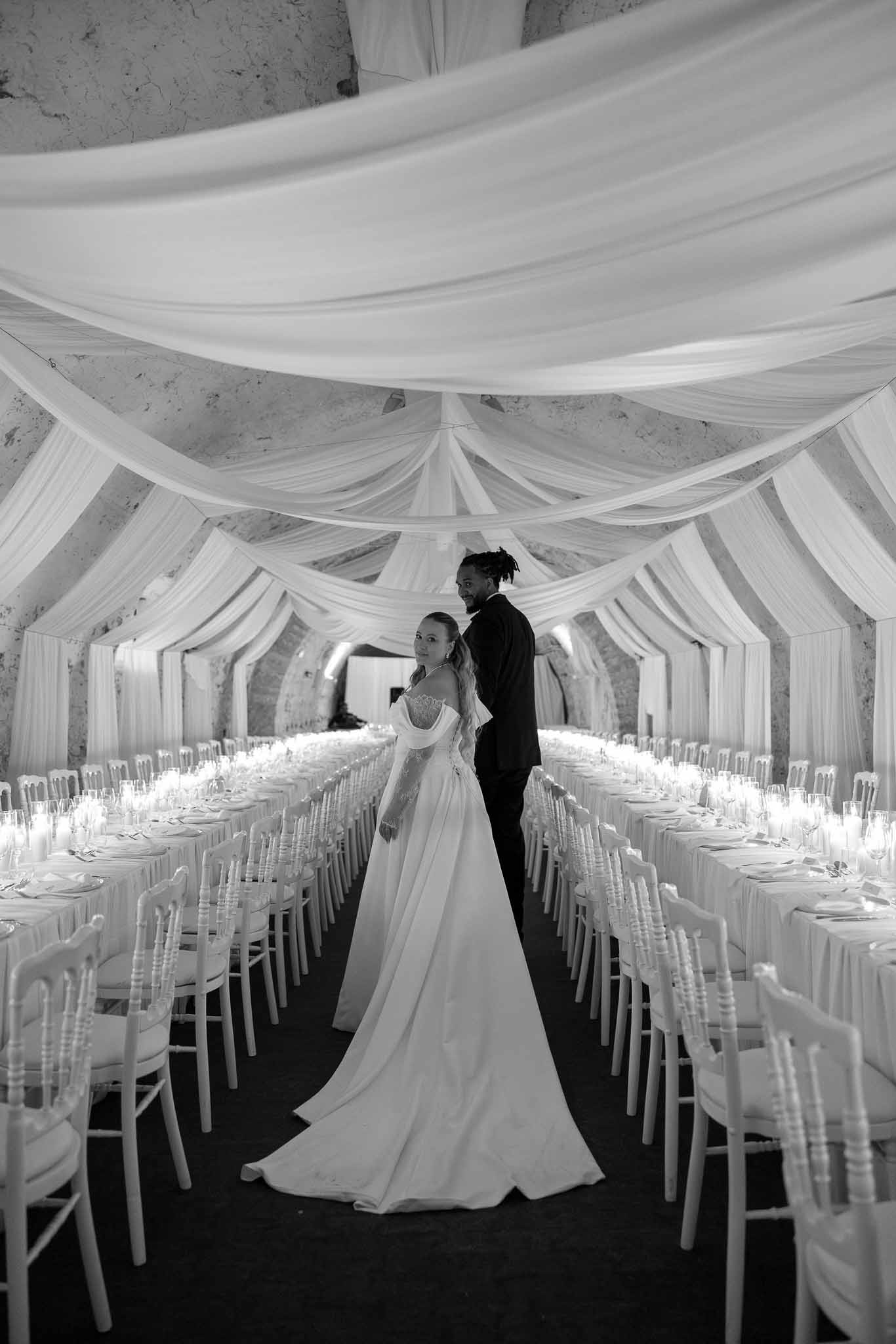 Black and white couple looking back between candlelit tables in vaulted stone hall with white draping